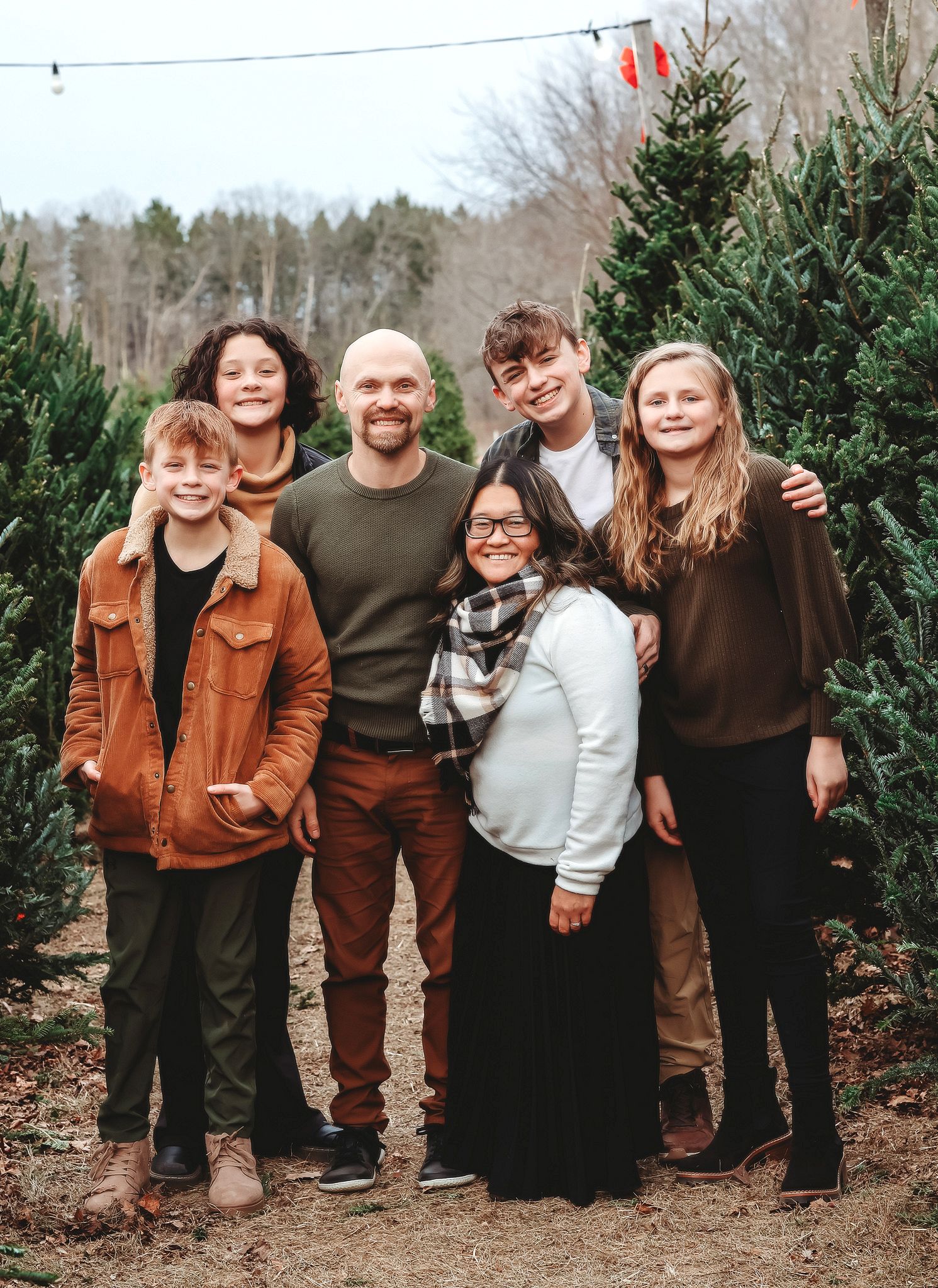 A bald father poses joyfully with his five children among Christmas trees outdoors.