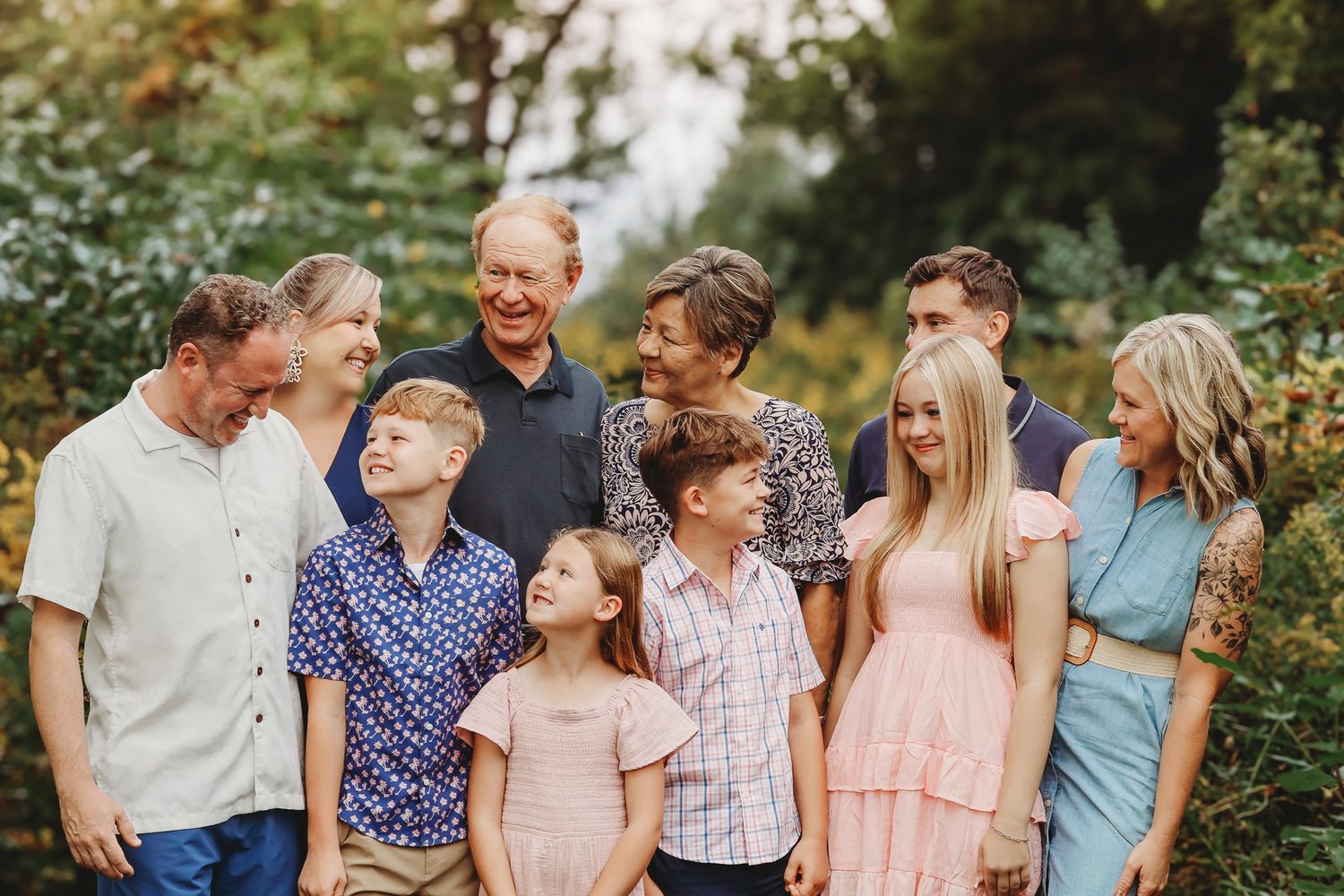 Large multigenerational family group smiling together outdoors surrounded by lush green trees.