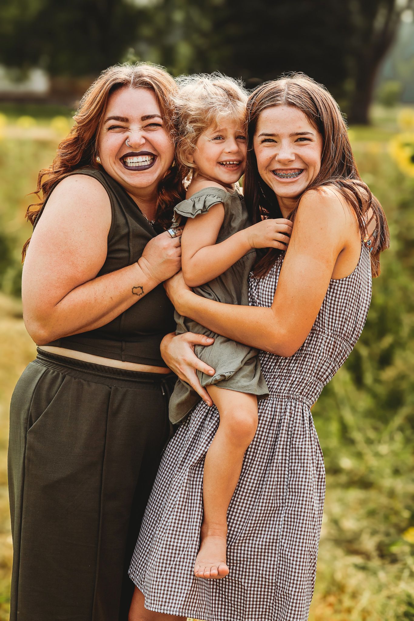 Three laughing females, two young women and a barefoot toddler, hugging outdoors in a field.