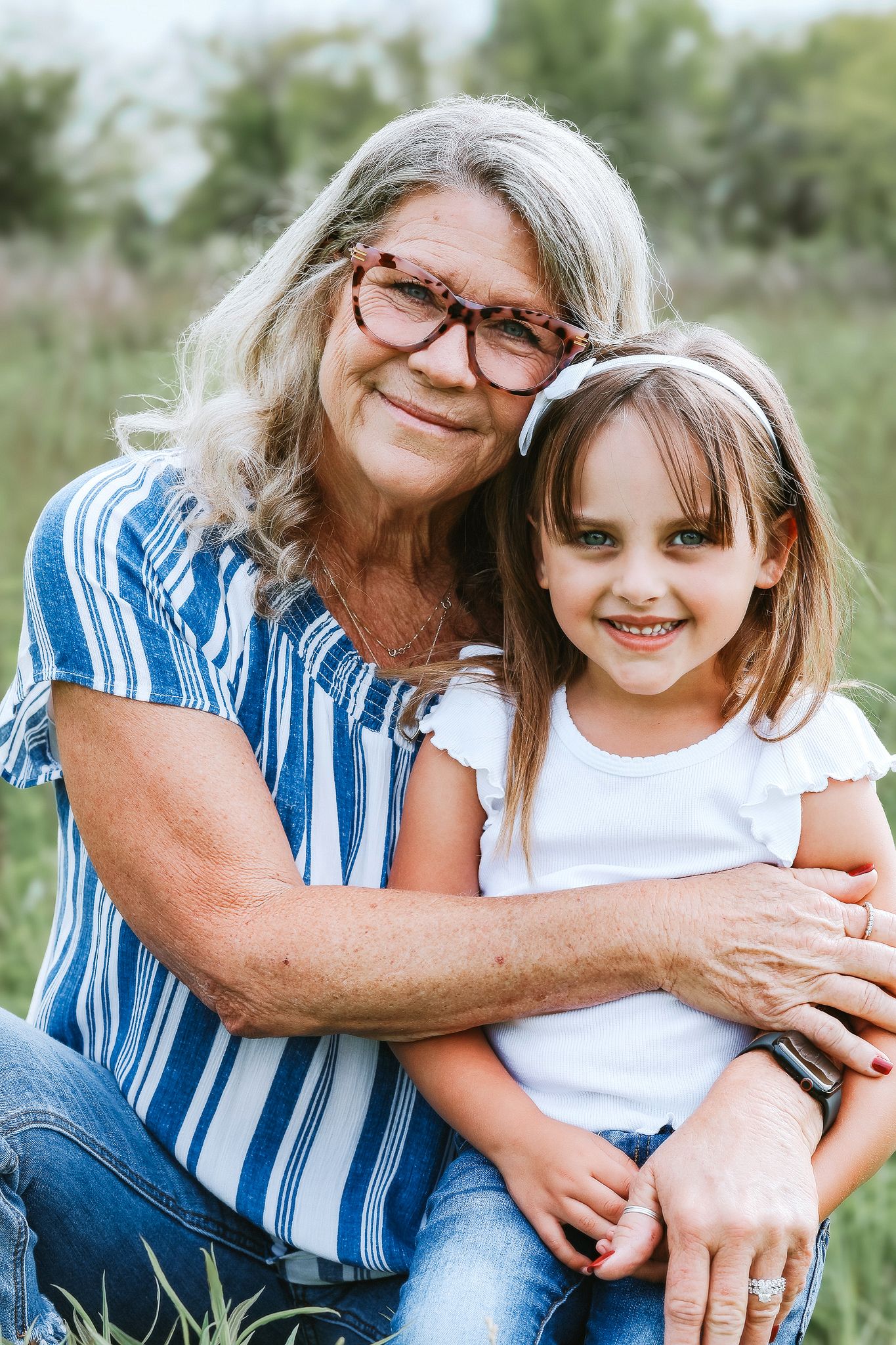Grandmother with glasses hugging young granddaughter outdoors in a grassy field.