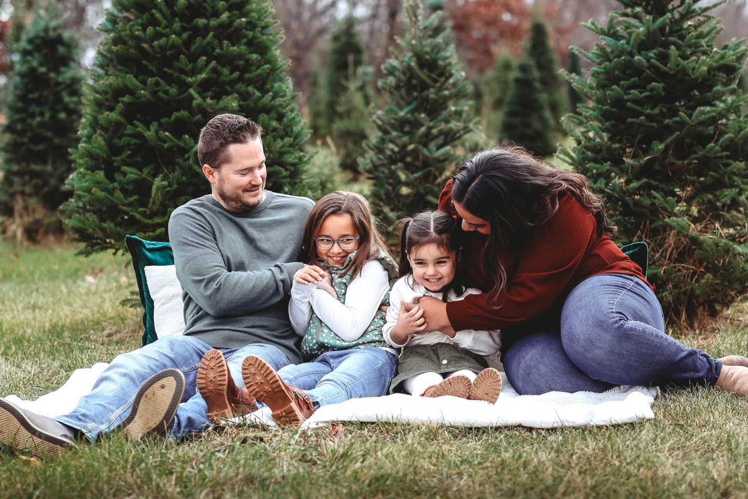 Loving family of four sits together on a blanket surrounded by Christmas trees.