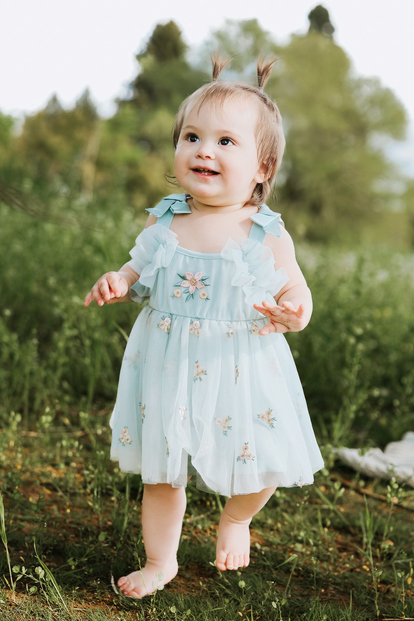 Smiling baby girl in light blue floral tulle dress standing barefoot outdoors in green grass.
