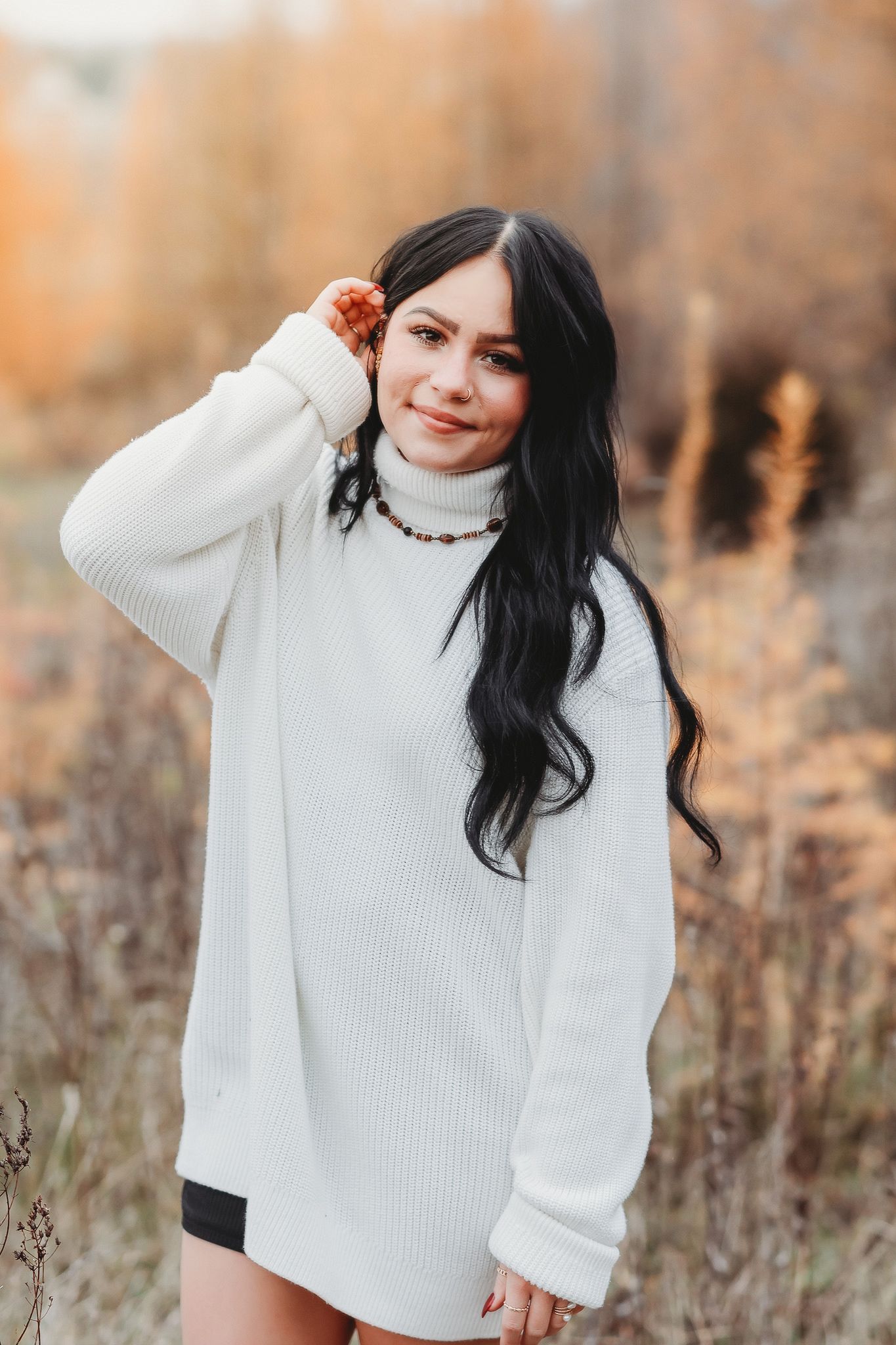 Young woman in oversized white knit sweater touching hair while posing in autumn nature setting.