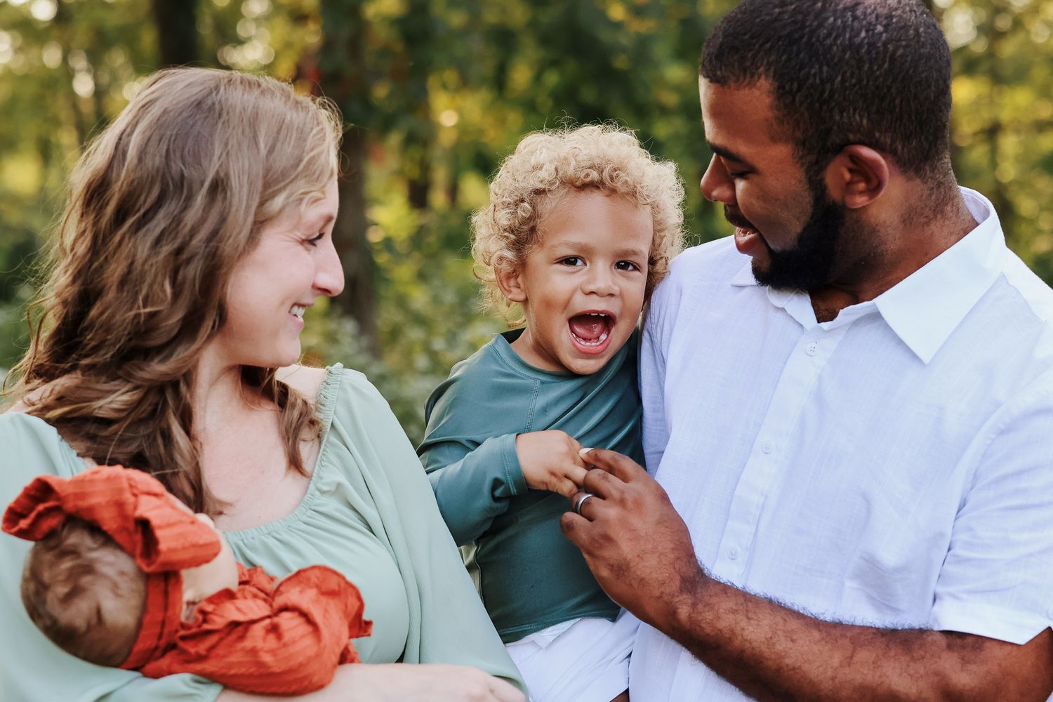Happy interracial family with newborn and toddler smiling outdoors.