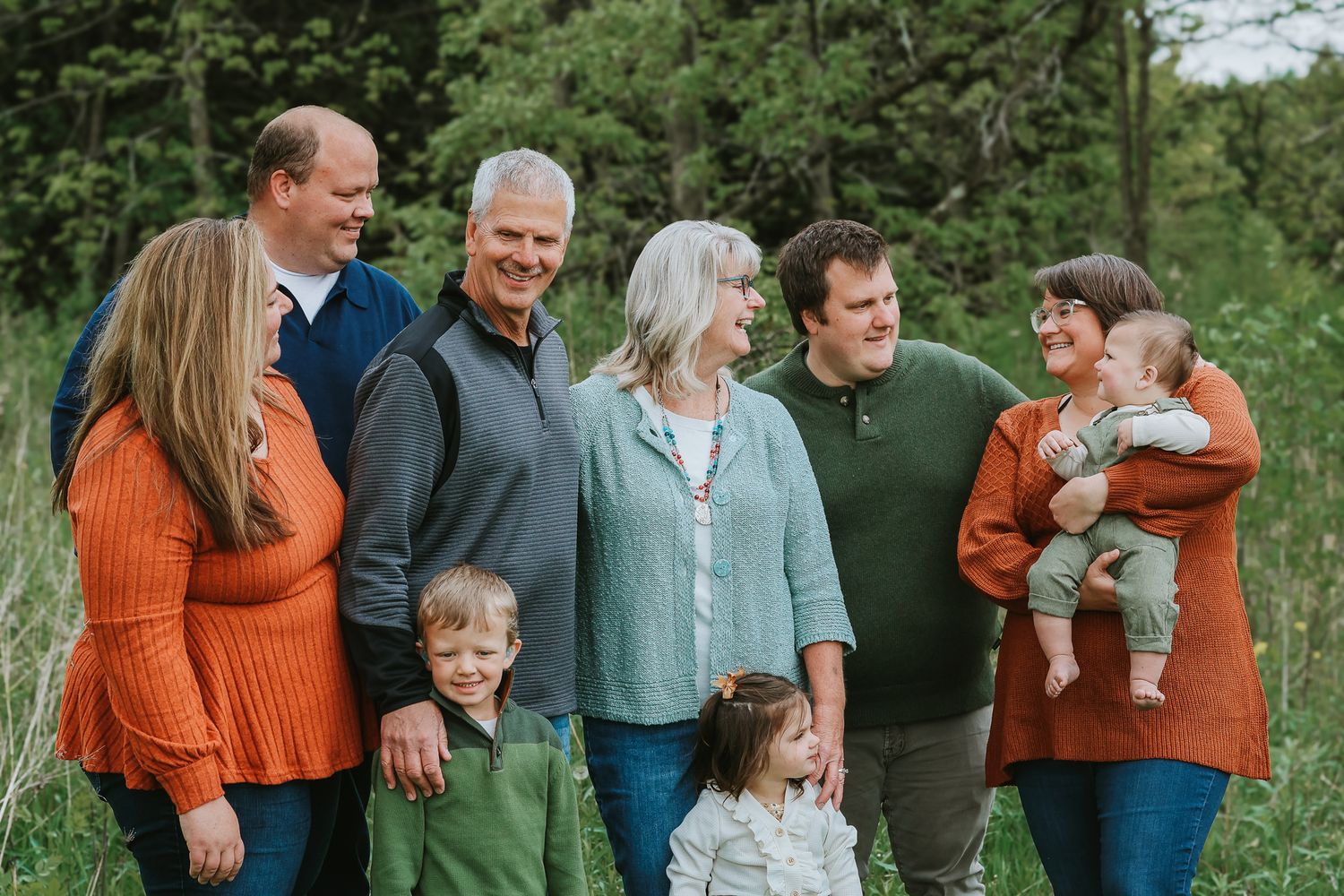A multigenerational family group smiles together outdoors in a lush green natural setting.
