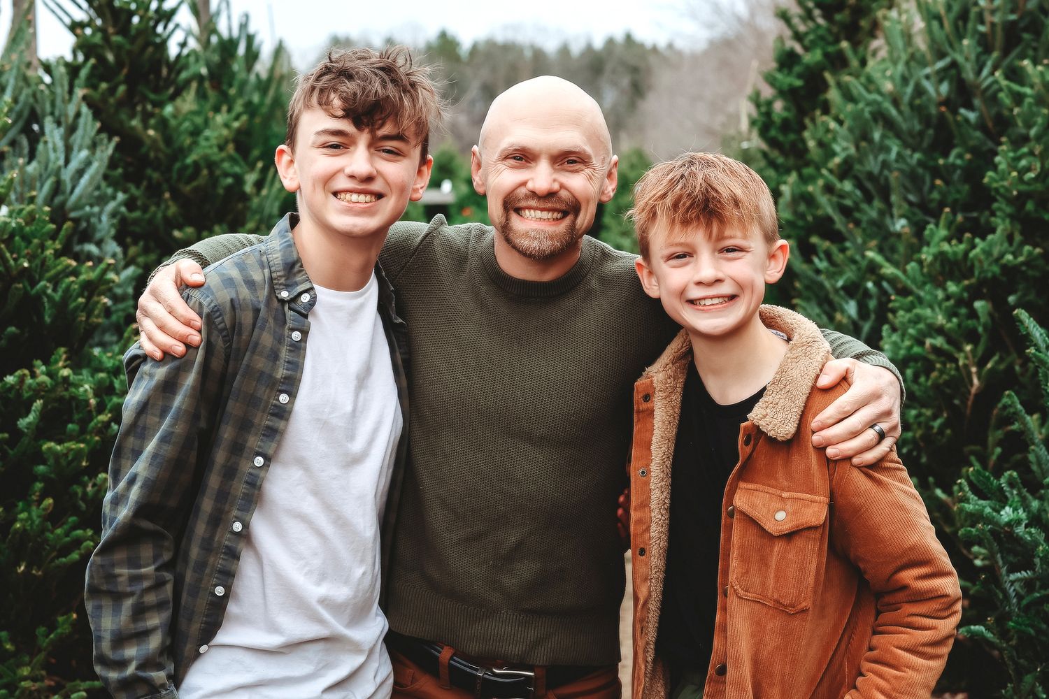 A bald father smiles proudly with his two teenage sons at a Christmas tree farm.
