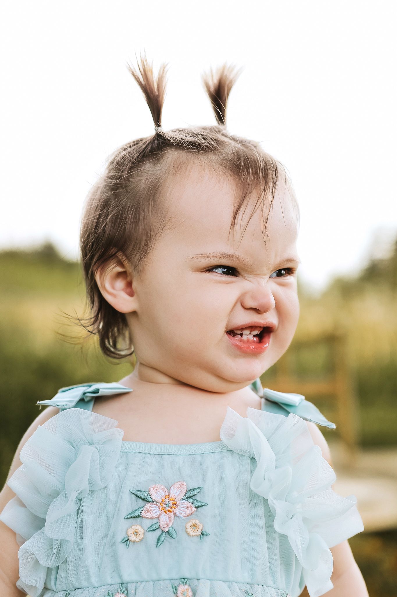 Expressive baby girl with tiny pigtails making a funny face in a light blue embroidered dress.