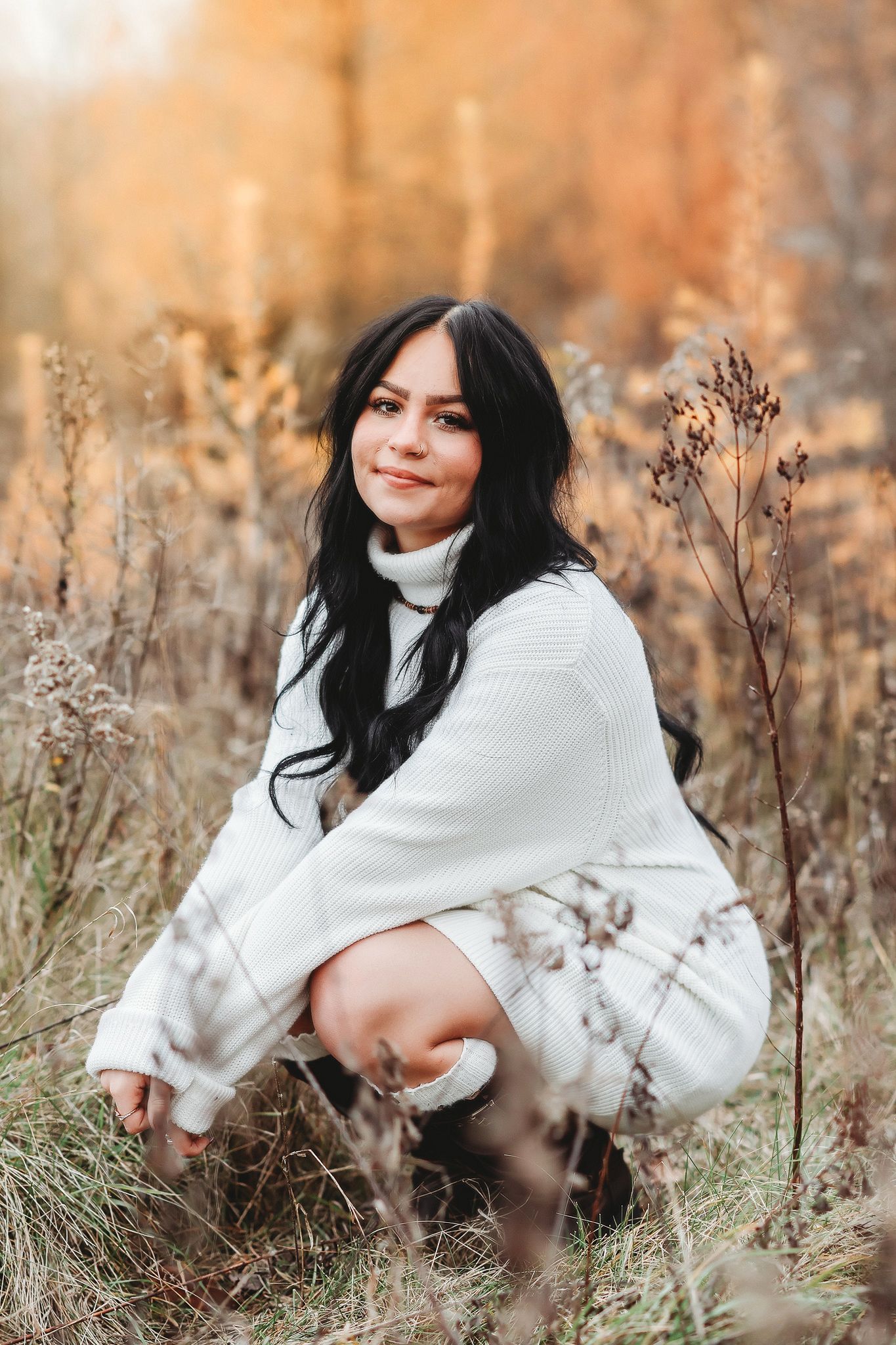 Dark-haired woman in white knit sweater crouching in autumn field with golden foliage background.
