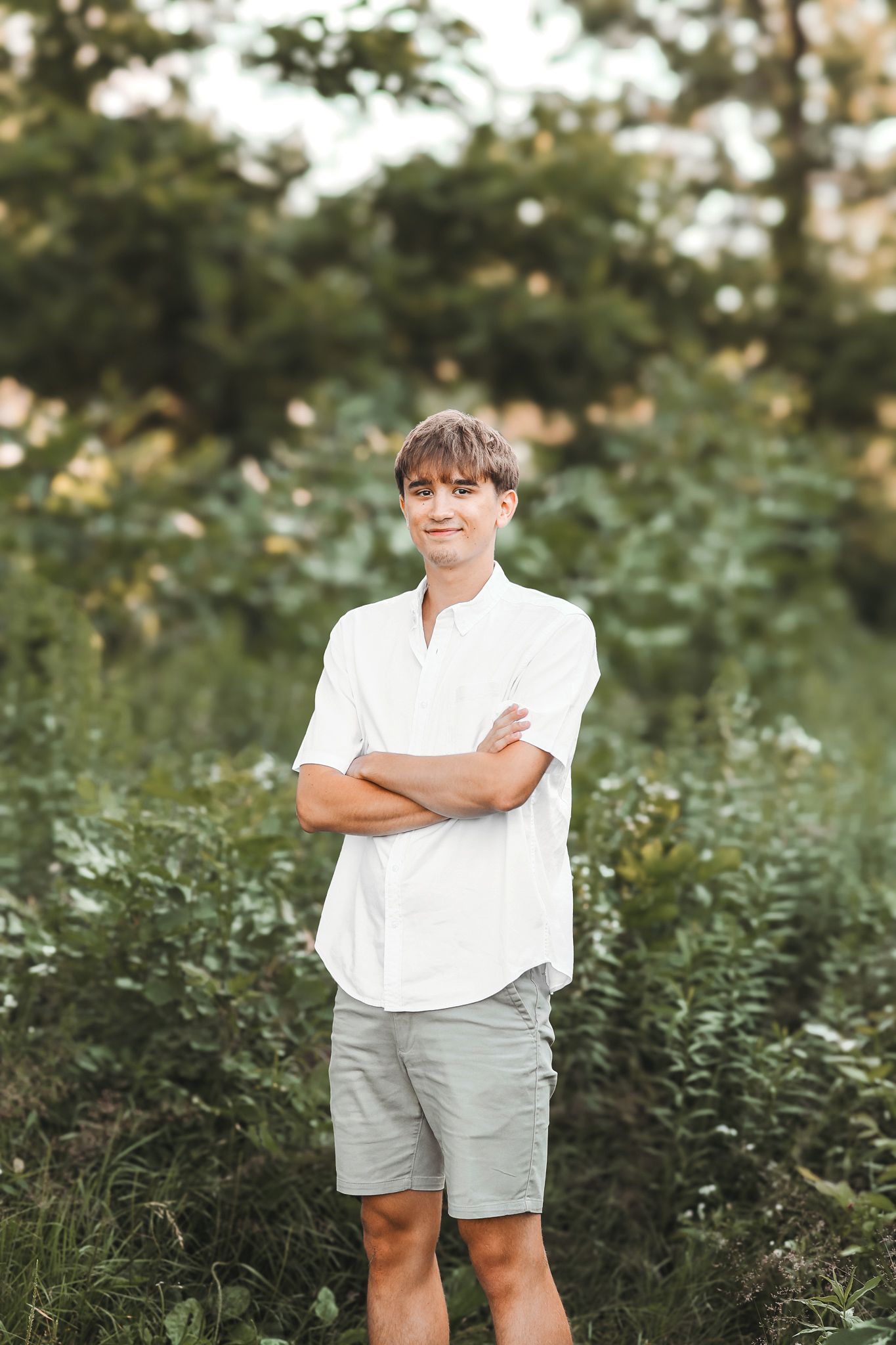 Young man in white shirt and gray shorts standing with arms crossed in a green outdoor setting.