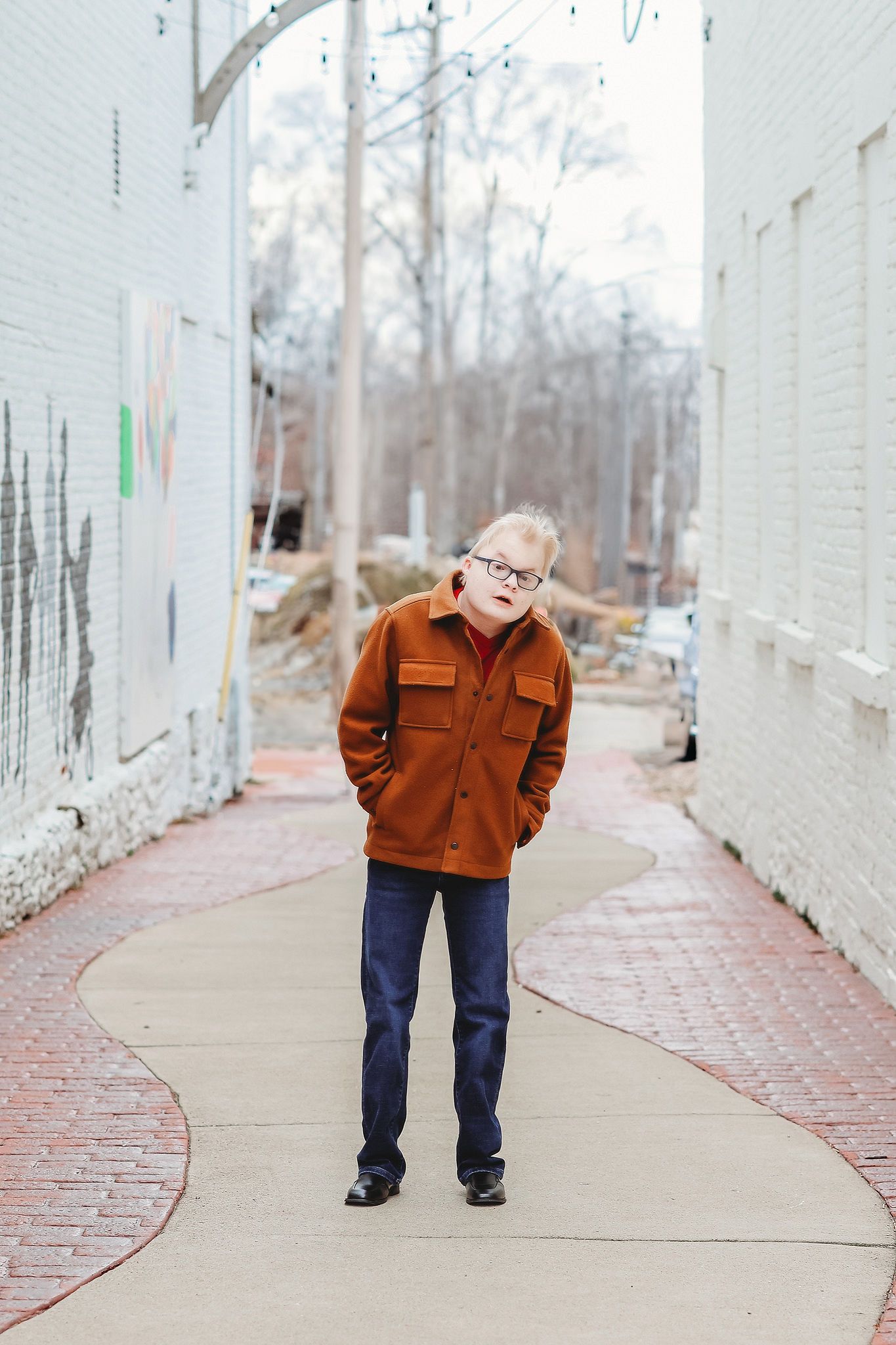 Young boy with Down syndrome in brown jacket and jeans standing on brick pathway between buildings.