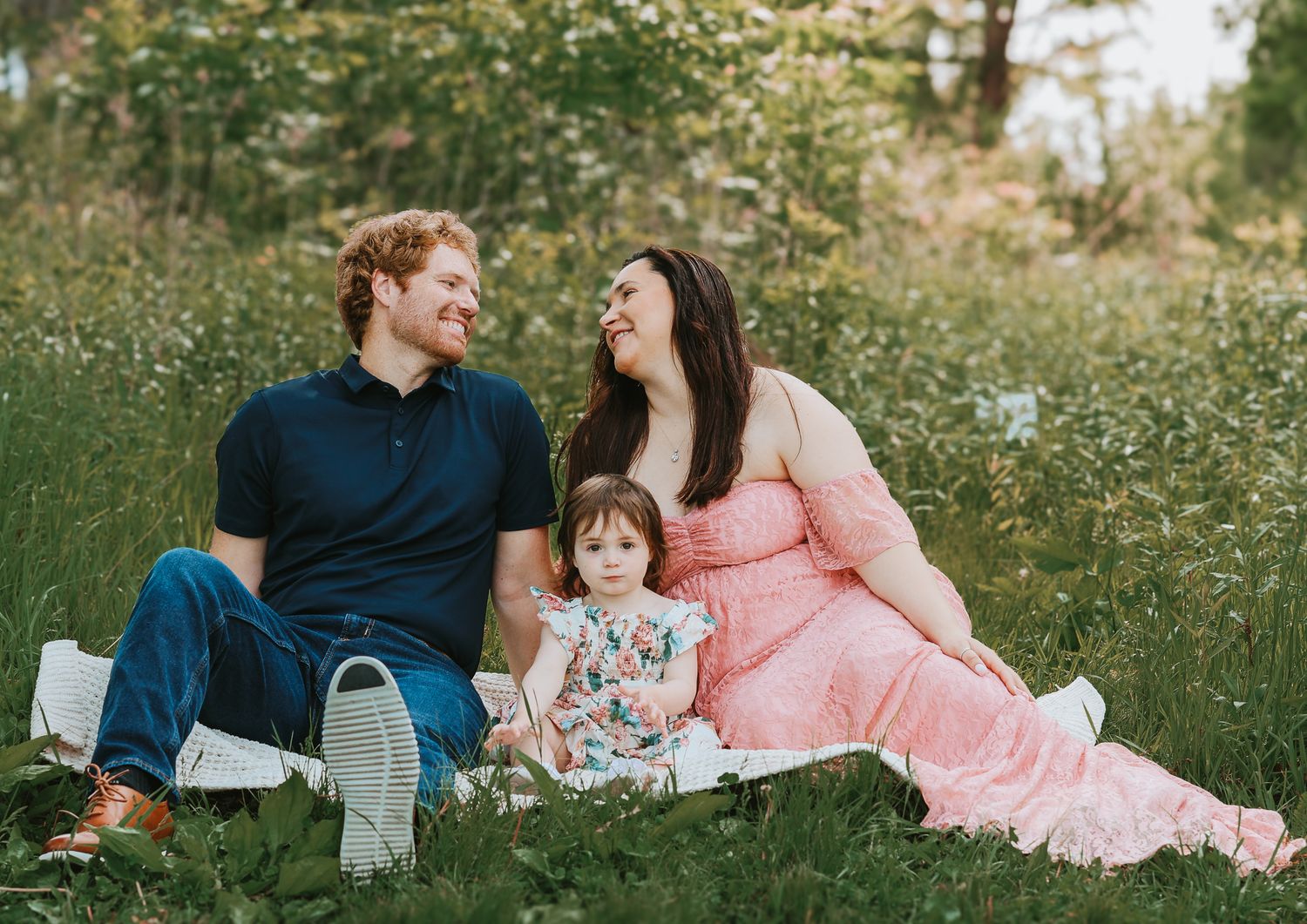 Joyful family portrait outdoors, redhead dad and pregnant mom in pink dress laughing with toddler on picnic blanket.