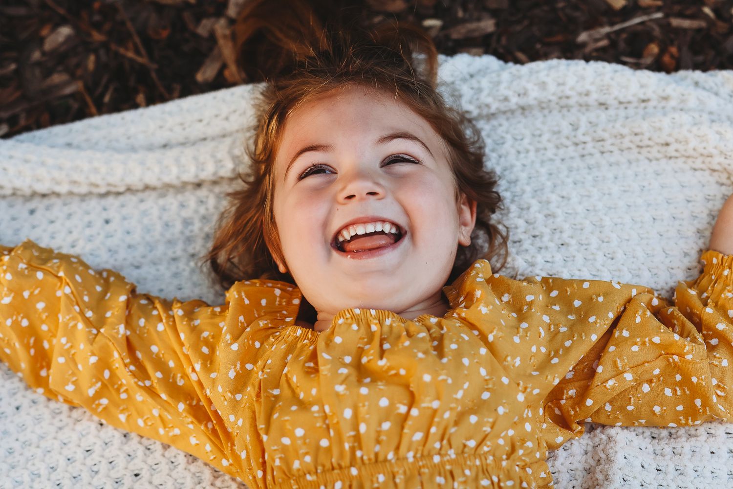 Happy young girl with red hair laughing while lying on a white knit blanket outdoors.