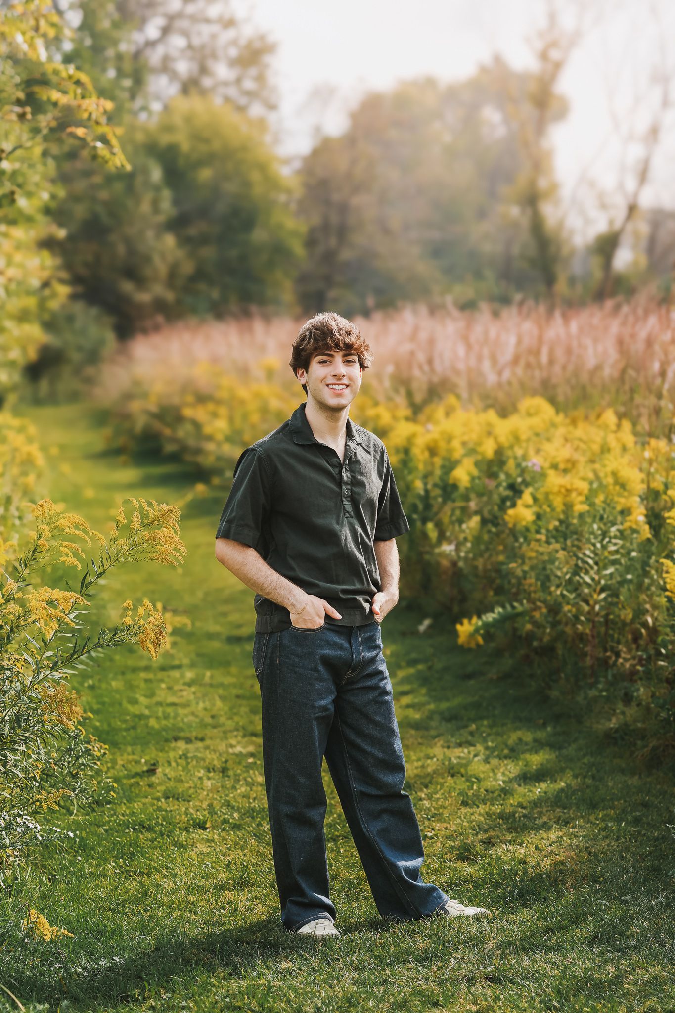 Smiling teenage boy in dark polo shirt standing on grass path surrounded by yellow wildflowers.