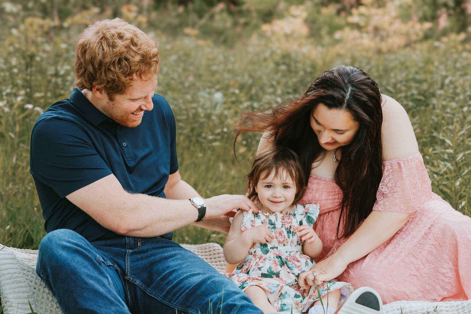 Happy family sitting in a wildflower meadow, parents smiling at their toddler daughter in a floral dress.