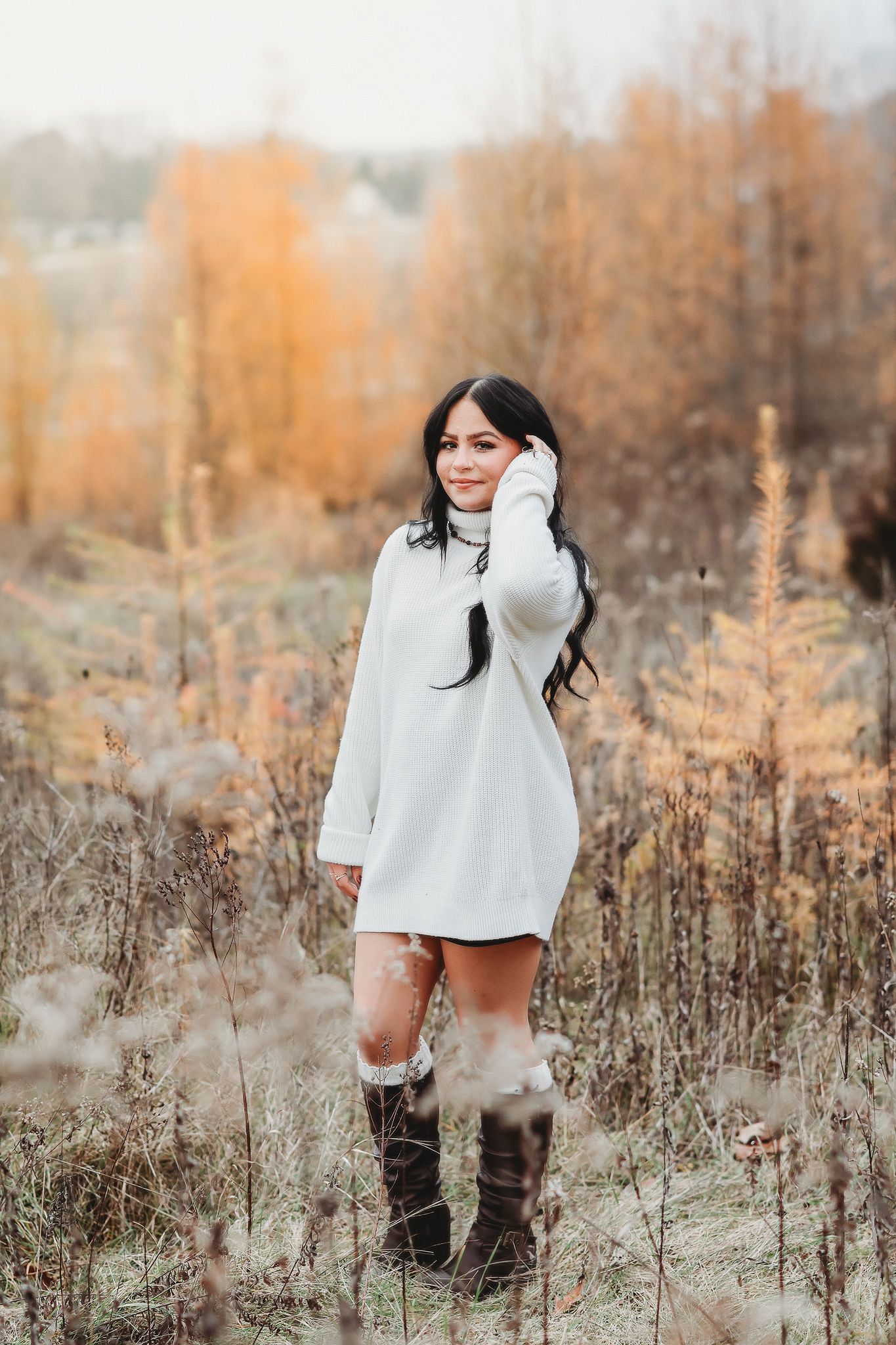 Young woman in white oversized sweater and tall brown boots standing in a misty autumn field.