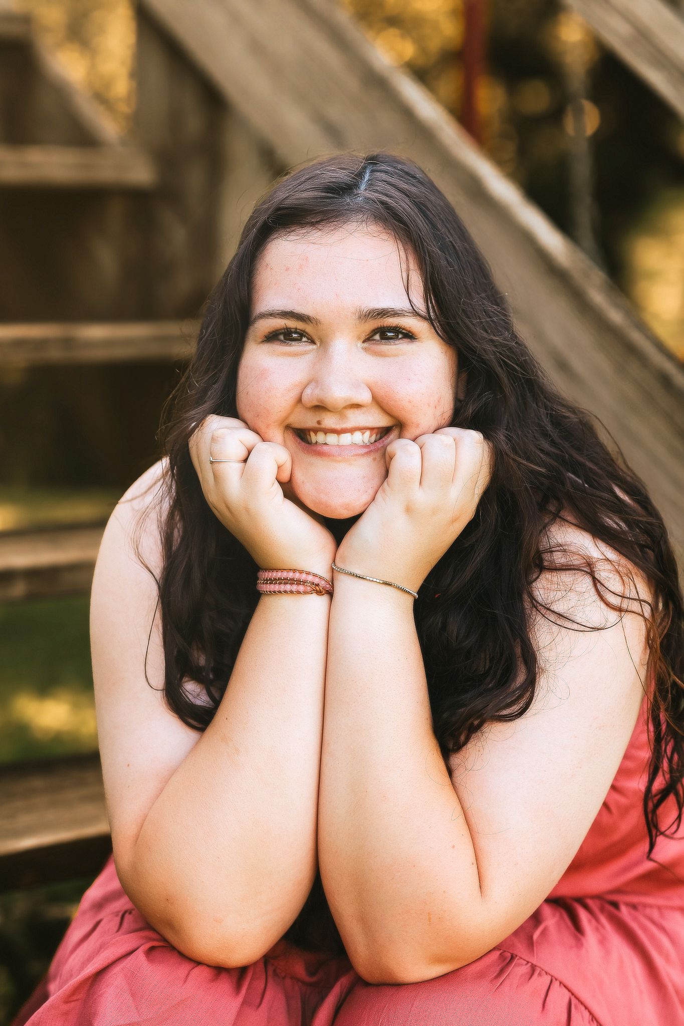 Cheerful young woman in pink dress resting chin on hands near wooden steps outdoors.