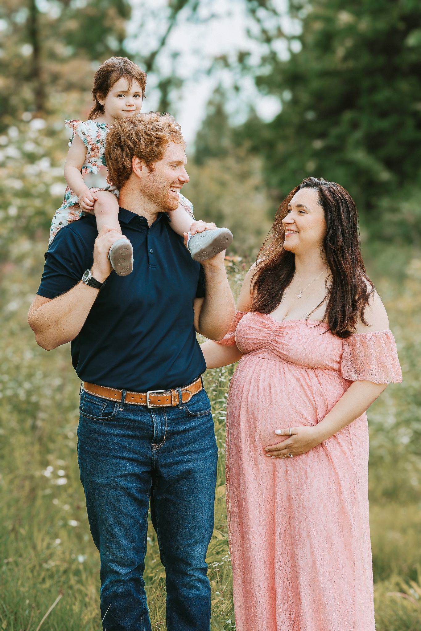 Father carrying toddler on shoulders smiling at pregnant wife in pink gown in a field.