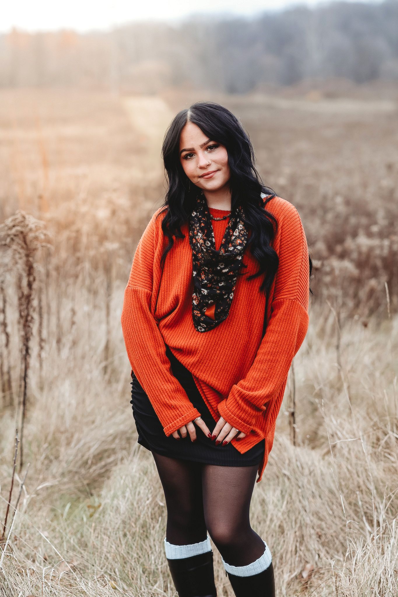 Young woman in oversized orange sweater and black skirt posing in a golden autumn field at sunset.
