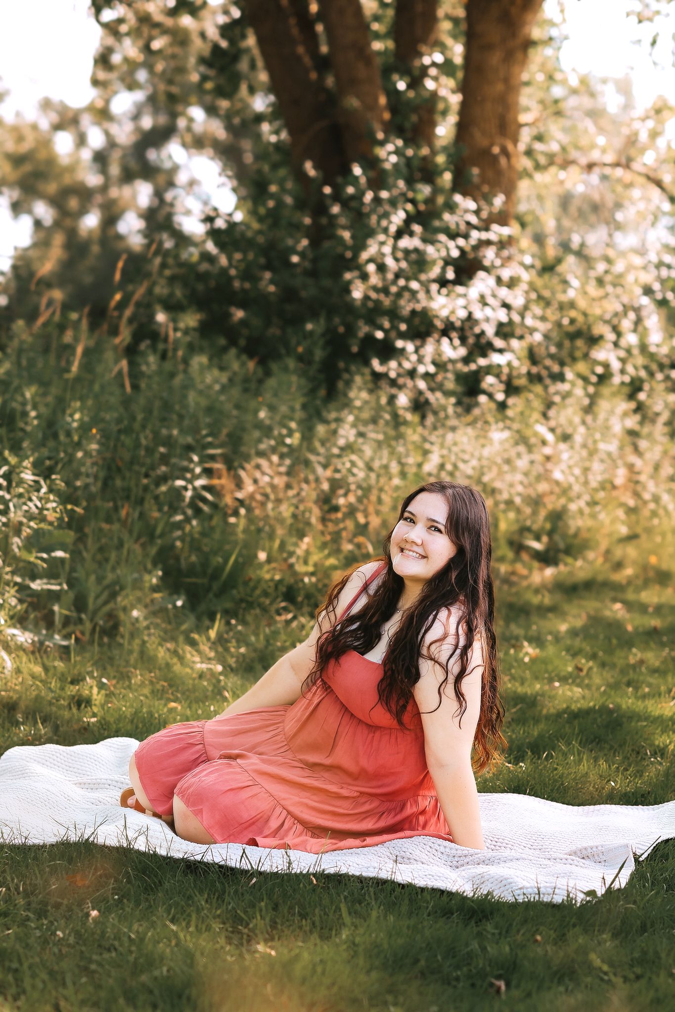 Young woman in pink dress sitting on blanket in sunny green meadow smiling.