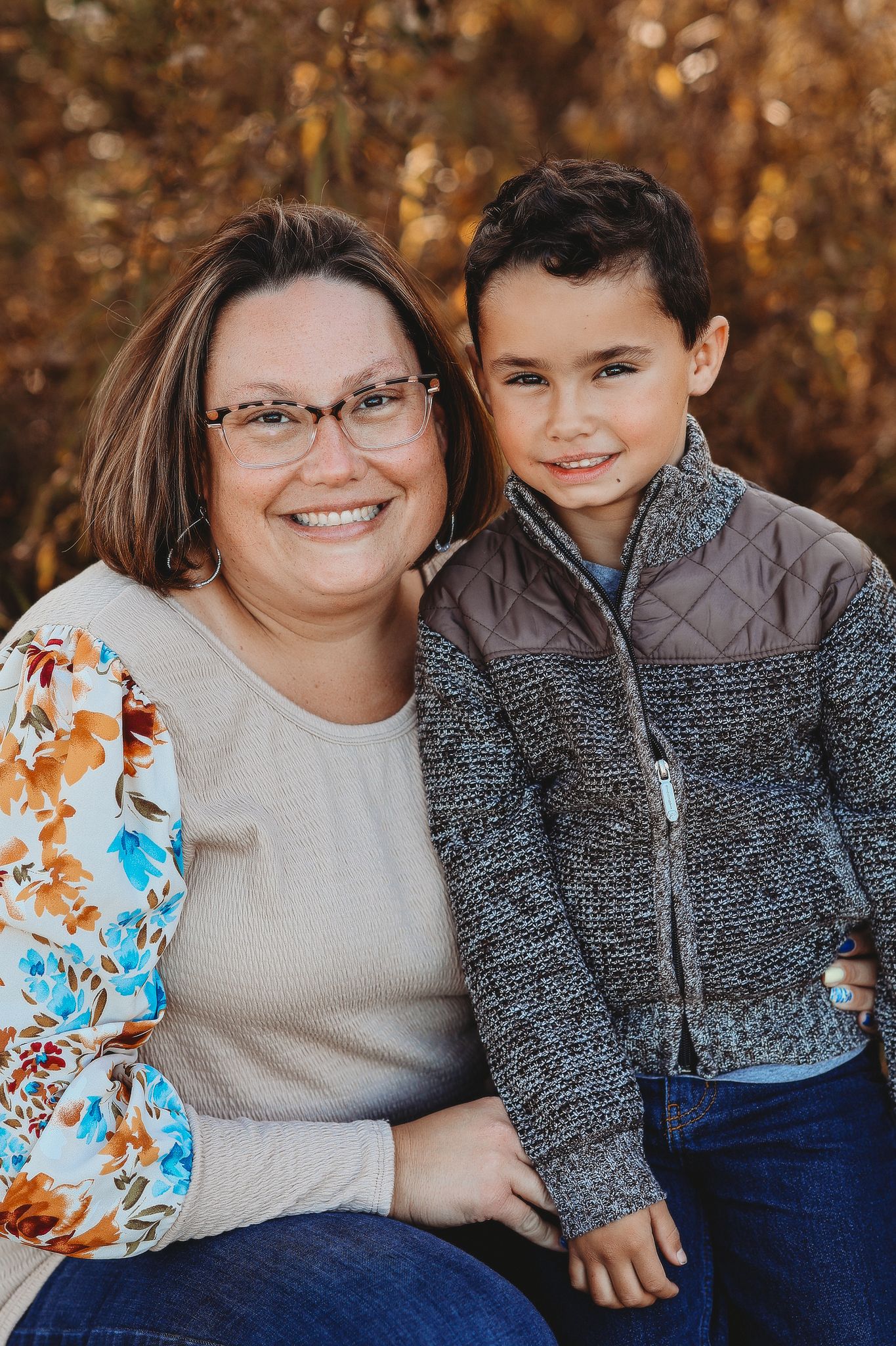 Smiling mother with glasses posing with her young son outdoors in autumn foliage.