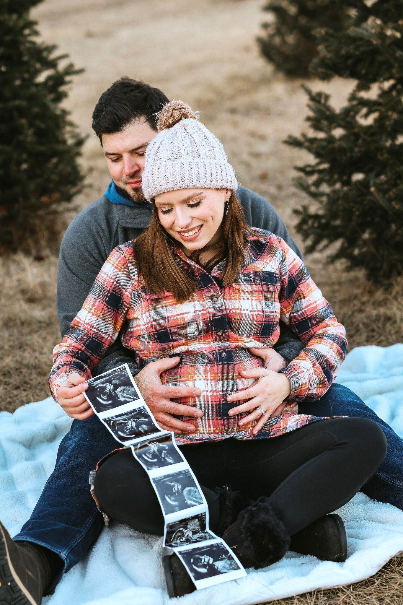 Couple sitting on blanket outdoors, cradling pregnant belly and holding ultrasound photos together.