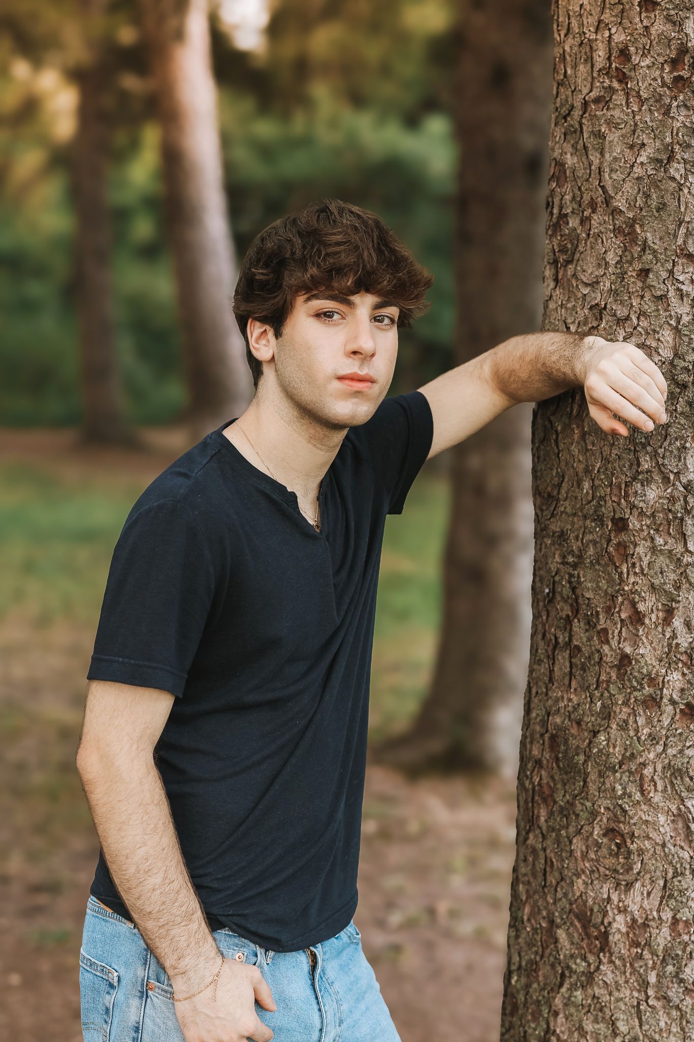 Young man in dark t-shirt leaning against tree trunk in sunlit wooded park setting.