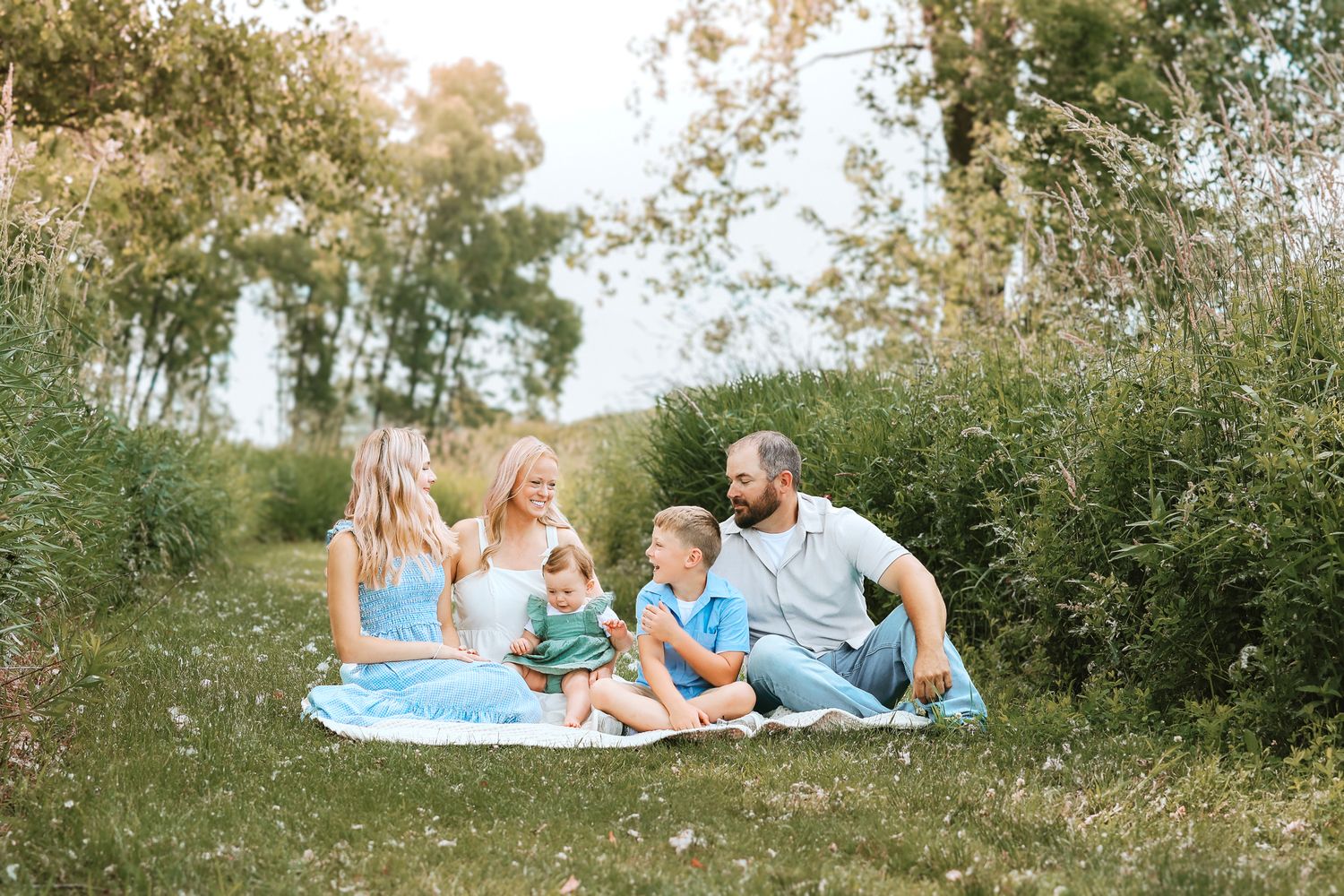 Family of five sitting together on a blanket in a green meadow, laughing and enjoying the outdoors.