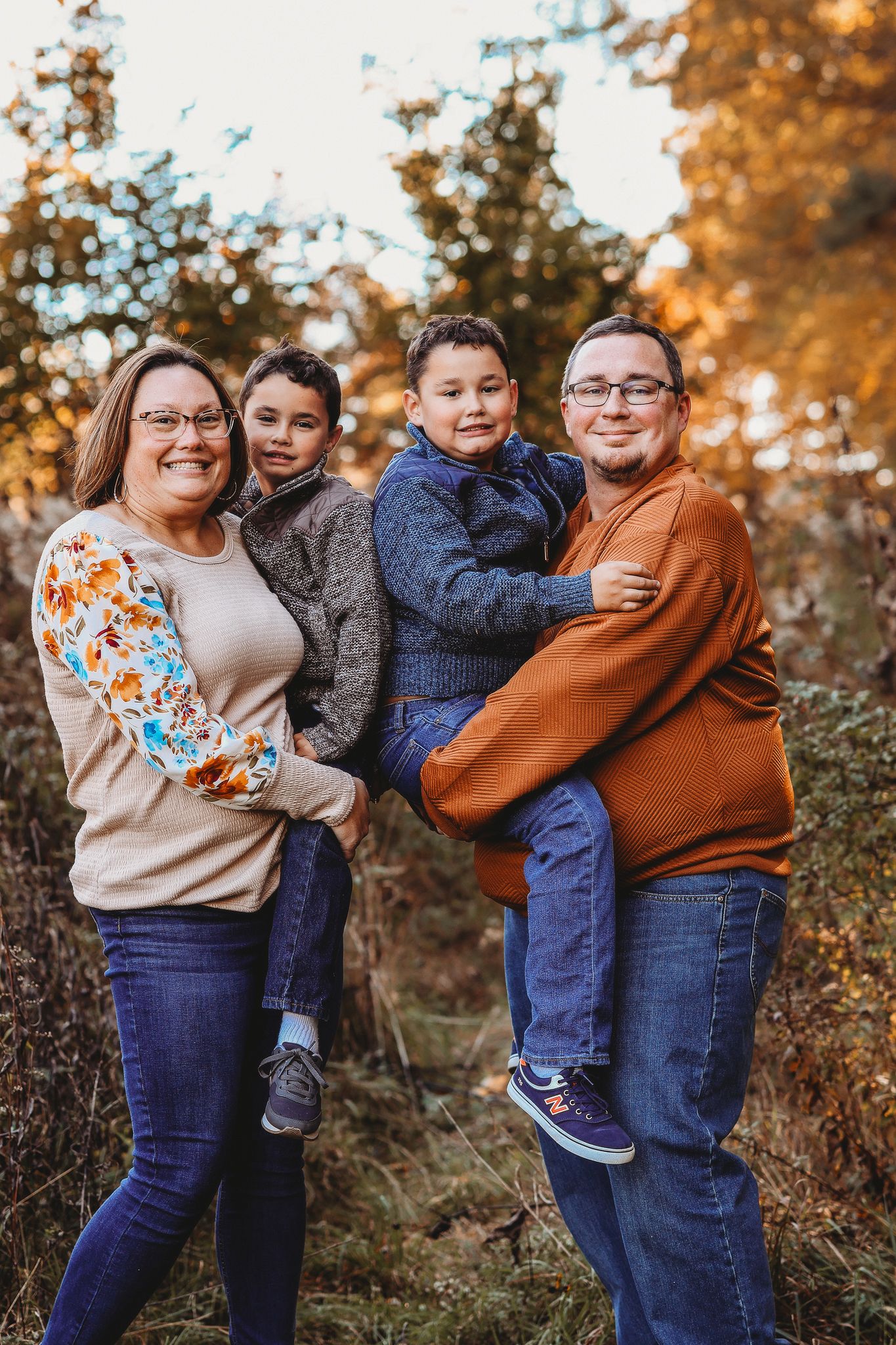 Family of four posing outdoors in fall foliage, parents holding two young boys, wearing autumn colors.