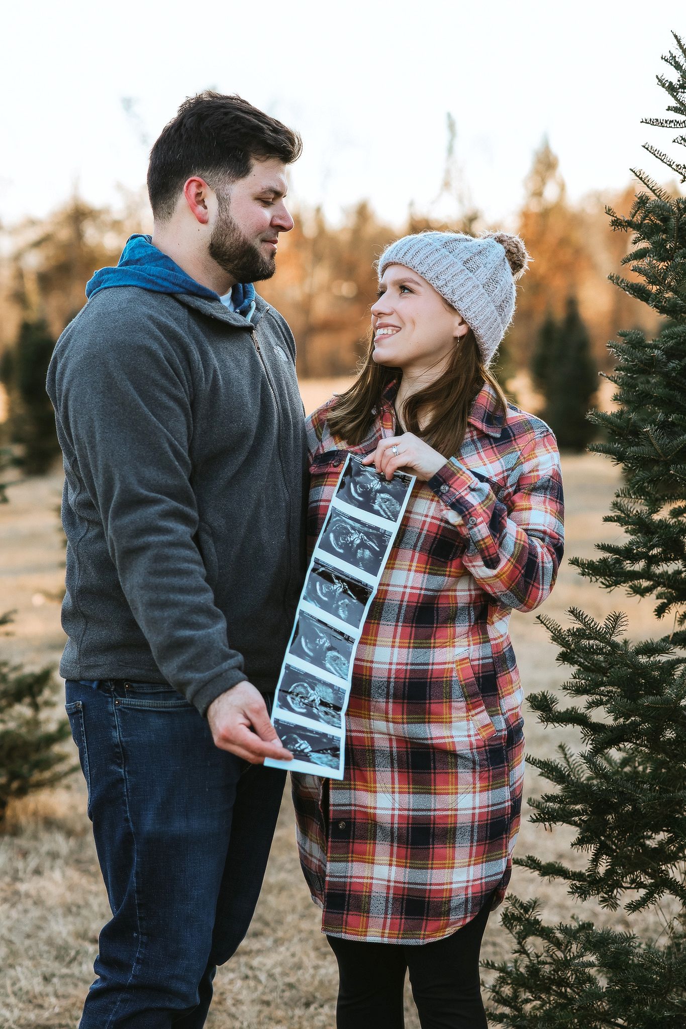 Couple sharing a tender moment outdoors holding ultrasound photos, announcing pregnancy near pine trees.