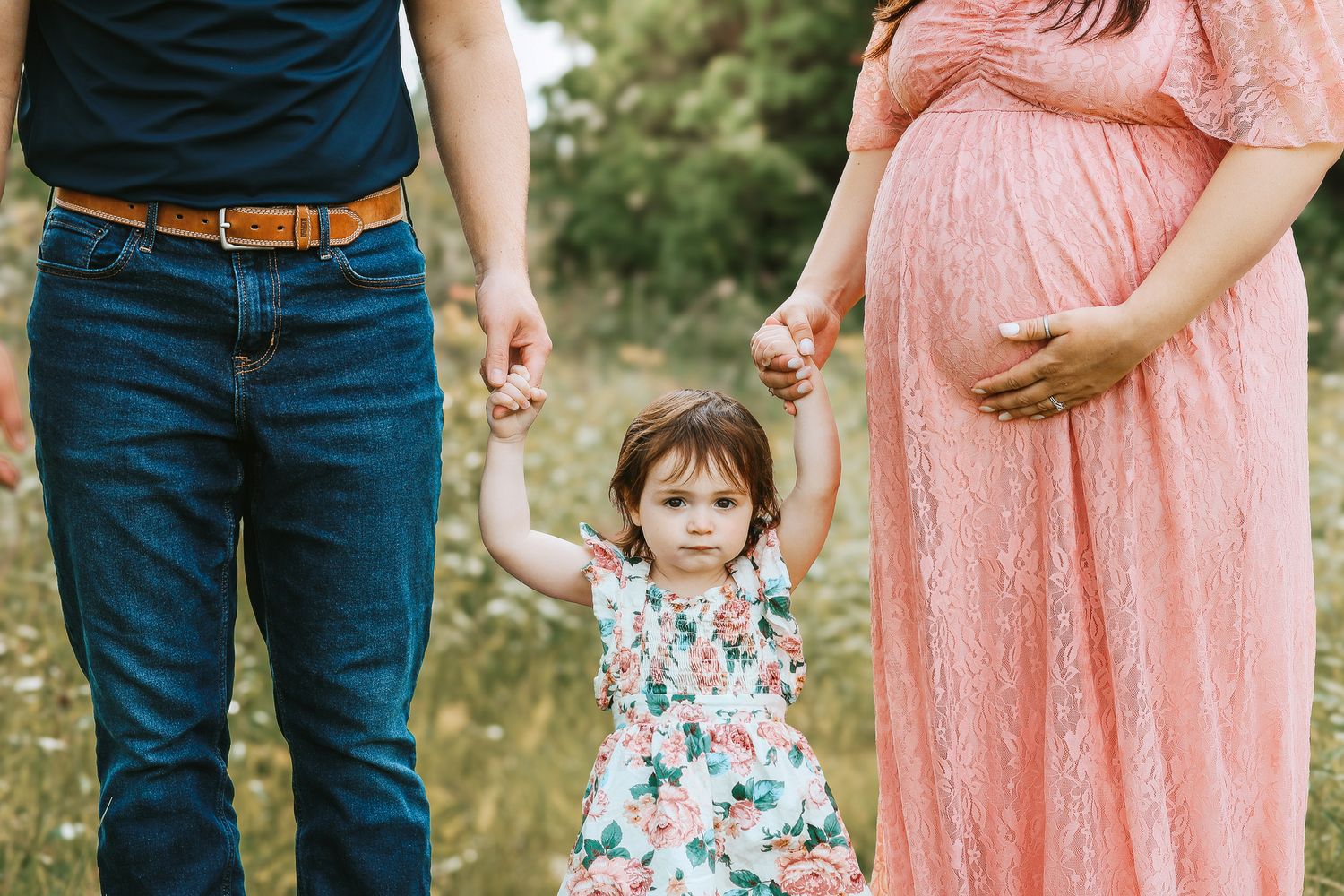 Toddler holding hands with parents in a sunny wildflower field during maternity shoot.