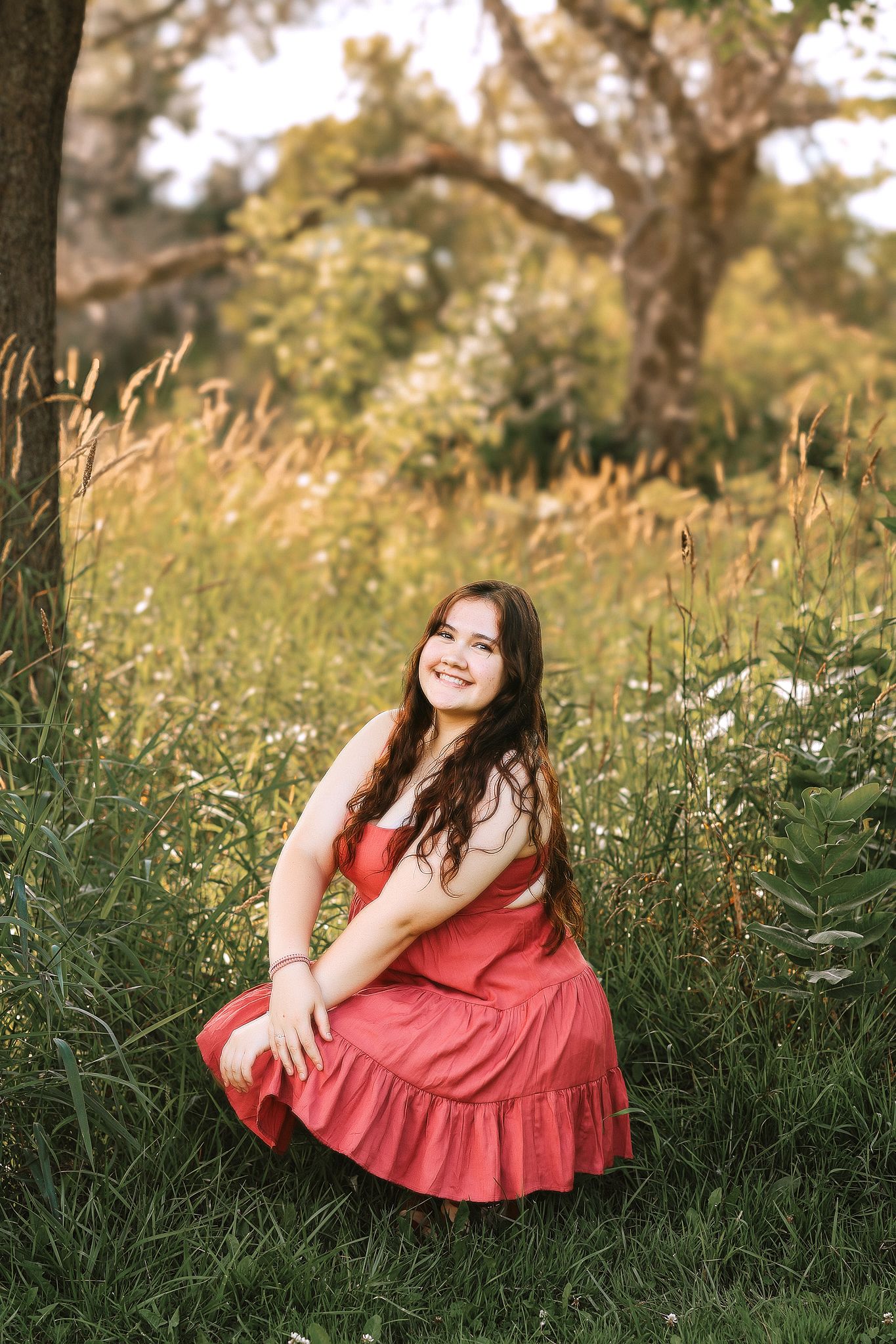 Smiling teenage girl in red dress sitting in tall grass field during golden hour sunlight.