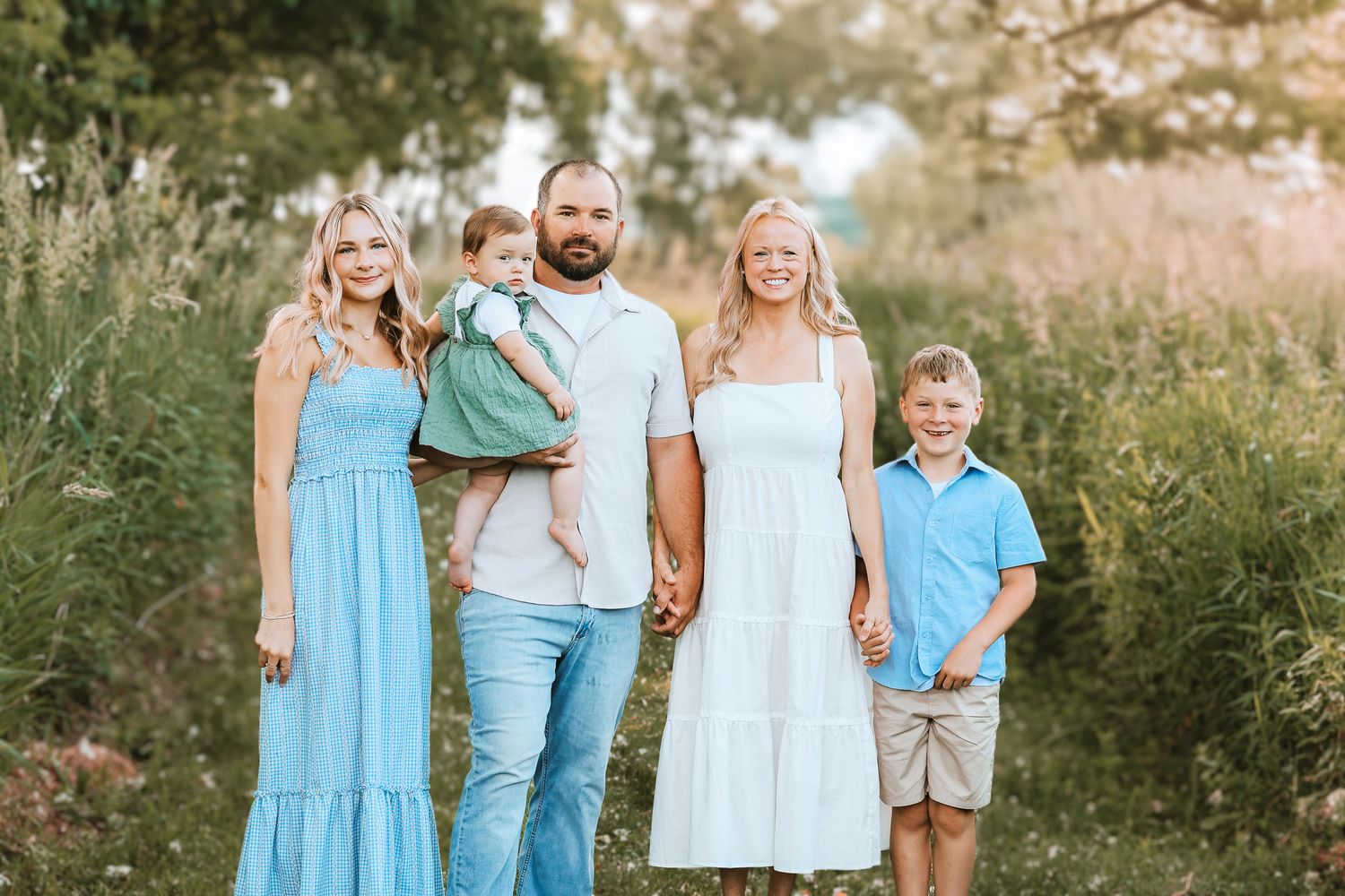 Smiling family of five holding hands in a sunlit meadow wearing coordinating blue and white summer outfits.