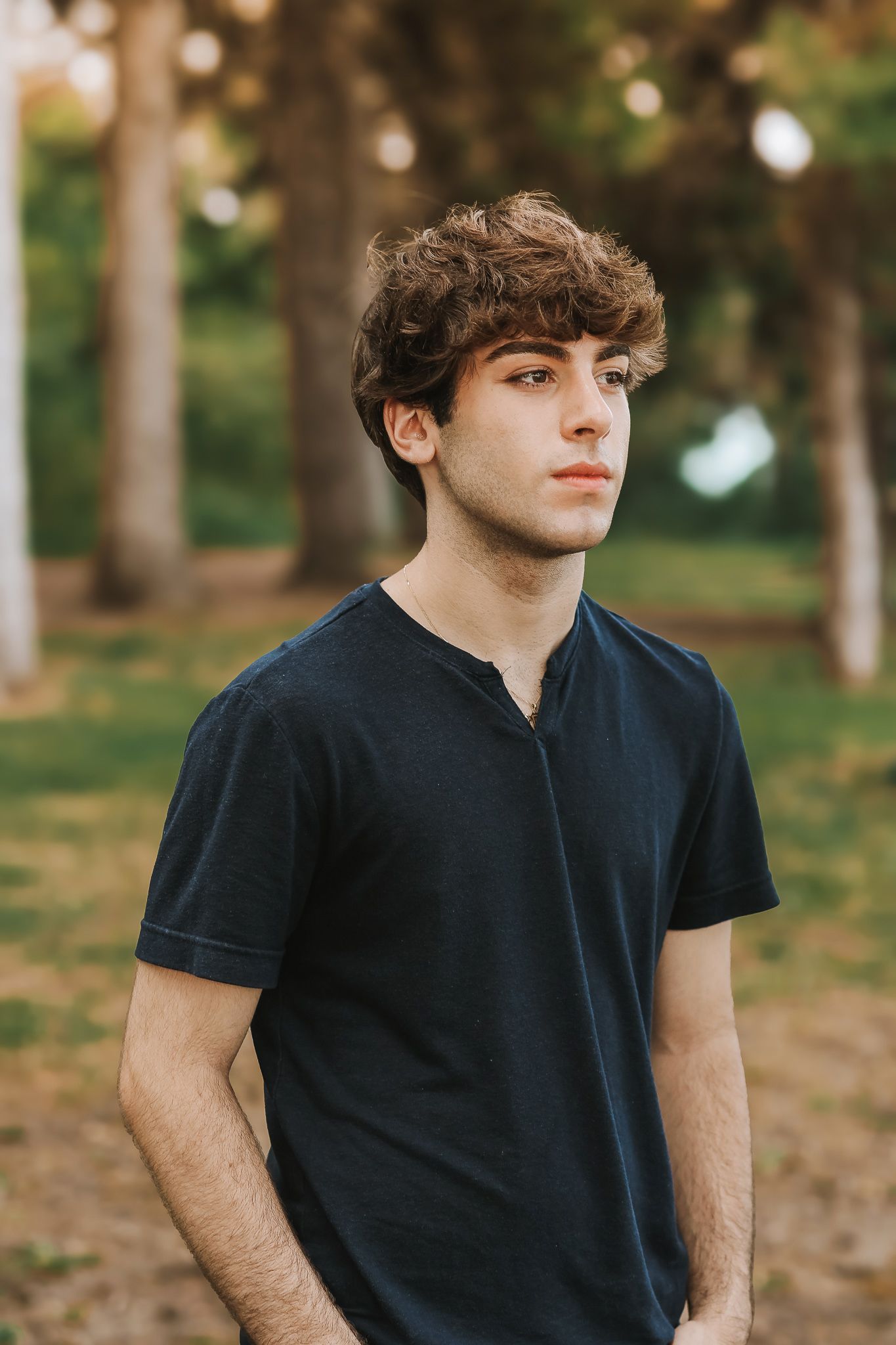 Teenage boy with curly hair wearing dark t-shirt posing outdoors in wooded park setting.