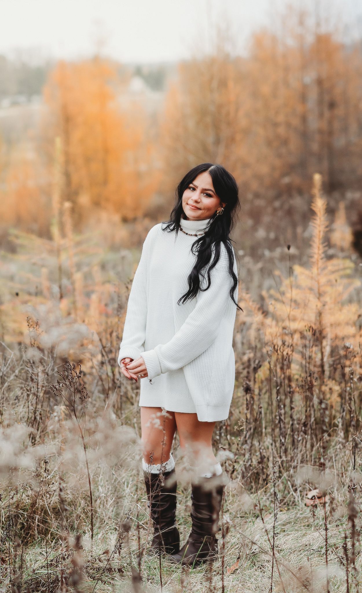 Beautiful brunette woman in white oversized sweater and tall boots poses in colorful fall foliage.