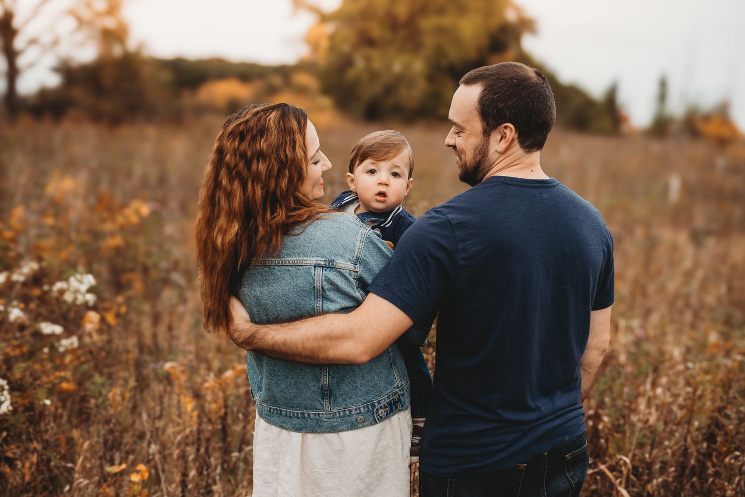 Young family stands together in a golden autumn field, parents gazing at their toddler.
