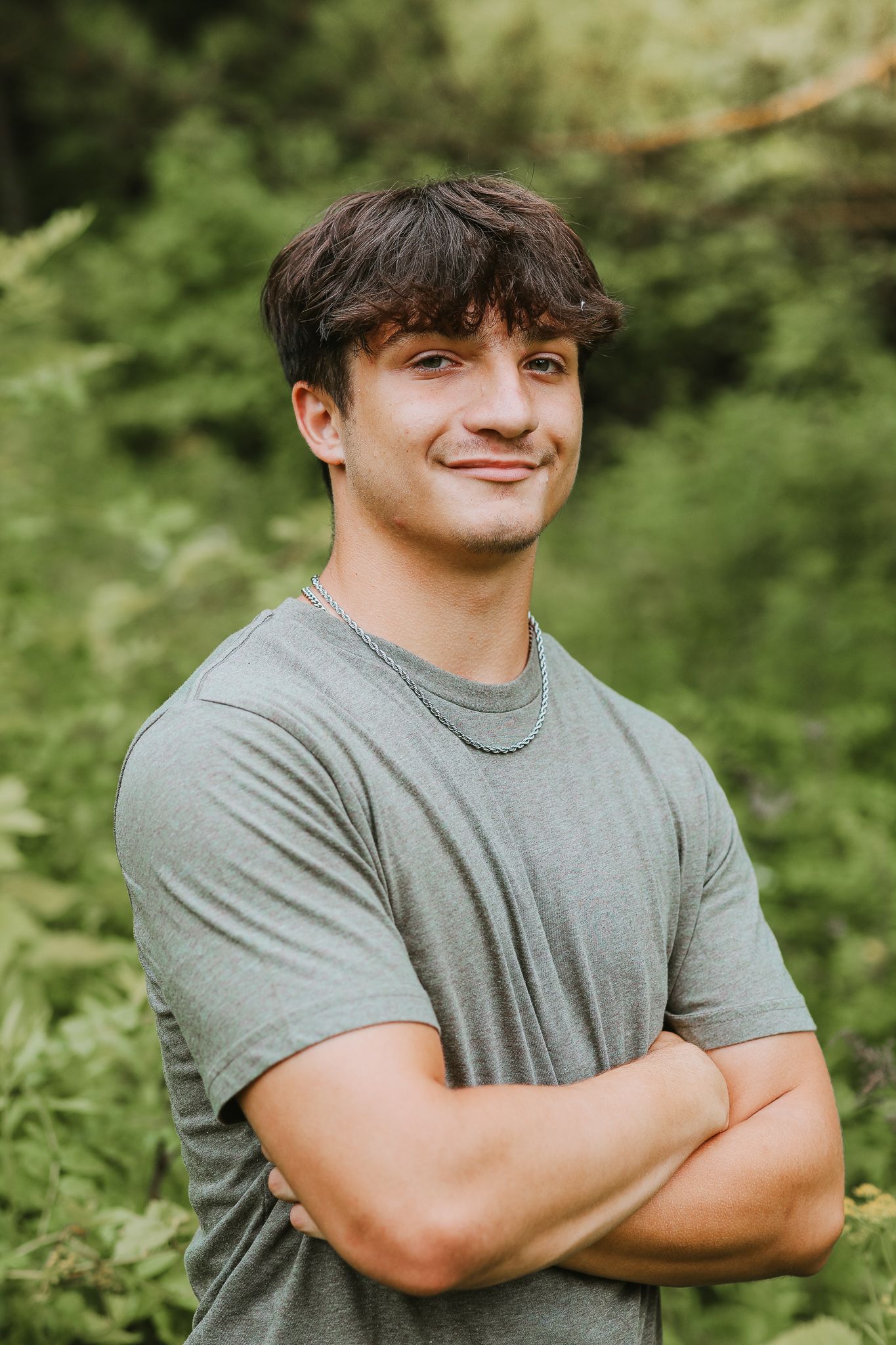 Young man in gray t-shirt with arms crossed smiling in lush green outdoor setting.