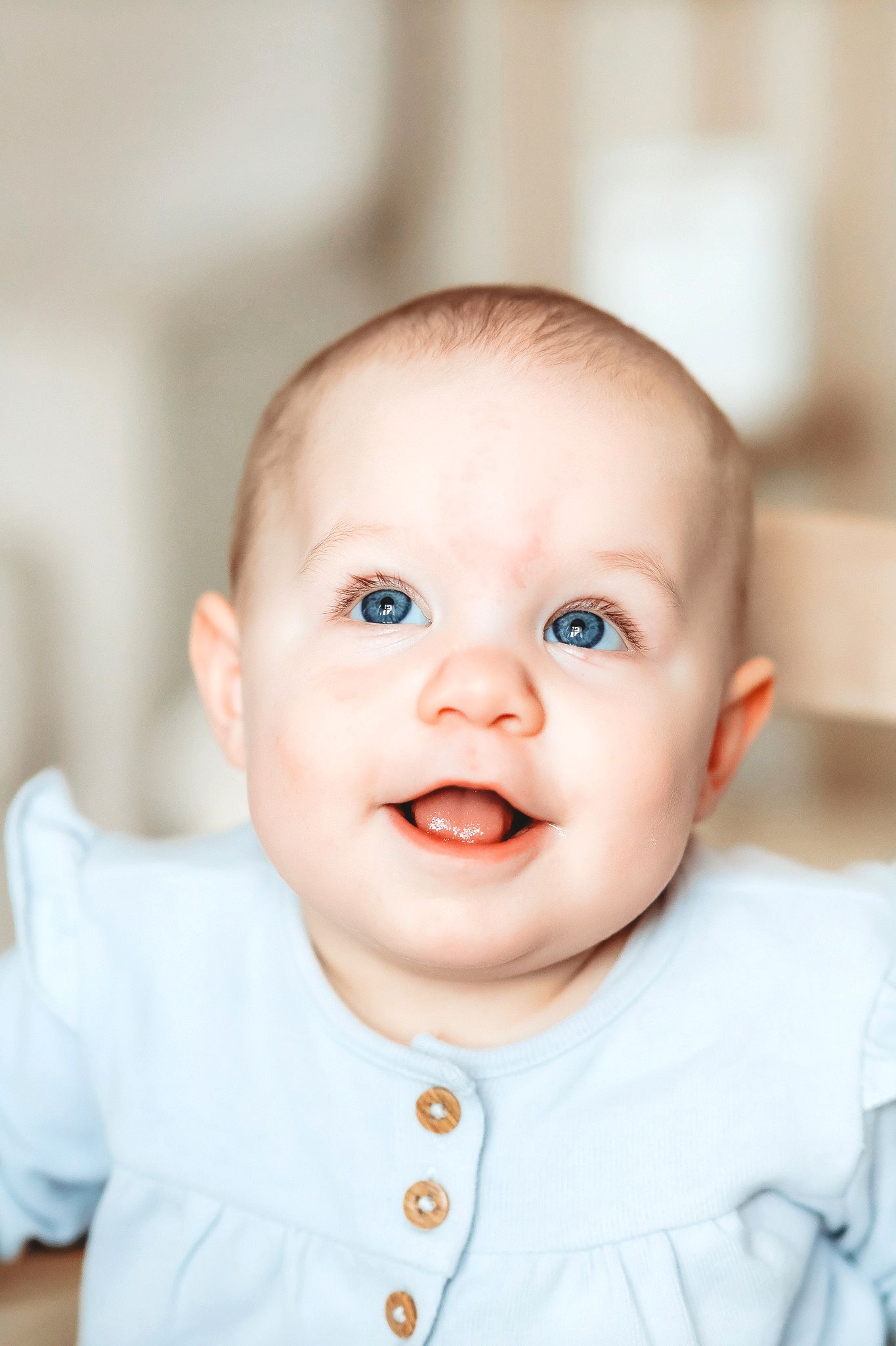 Adorable blue-eyed baby girl in a light blue button-up romper smiling brightly.