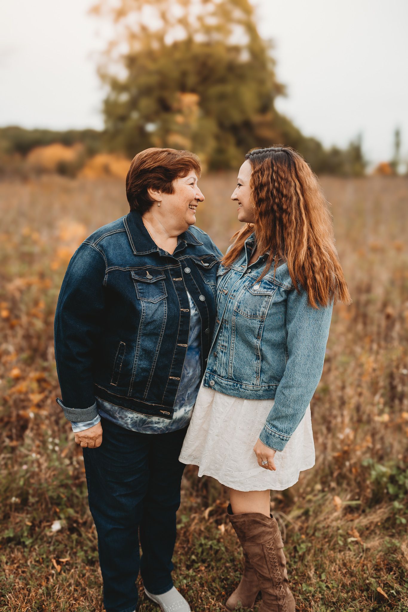 Two women in denim jackets smile at each other while holding hands in an autumn field.