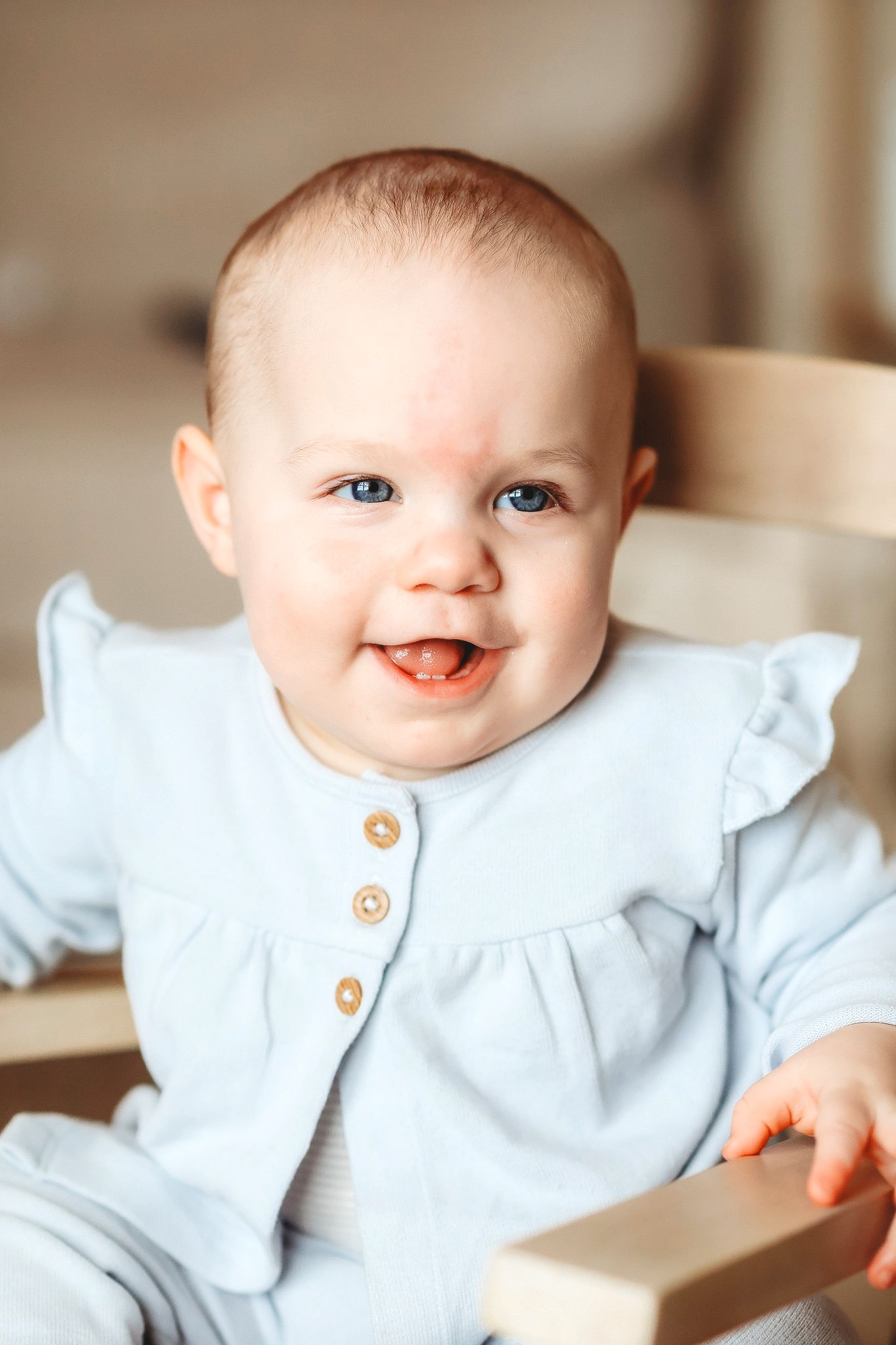 Happy blue-eyed baby girl in a light blue ruffled romper sitting in a wooden chair.