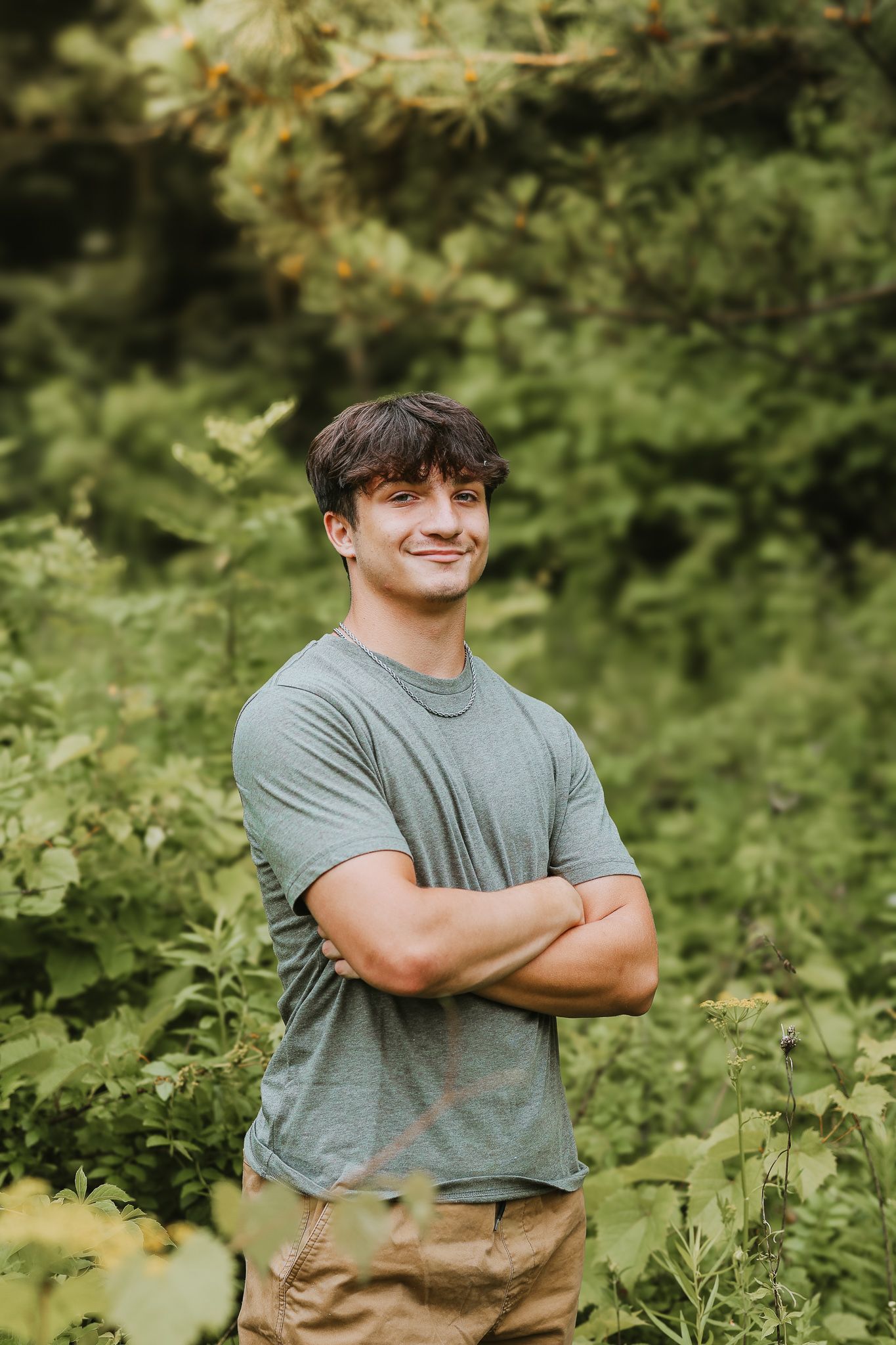 Confident young man in gray tee with arms crossed smiling in green forest setting.