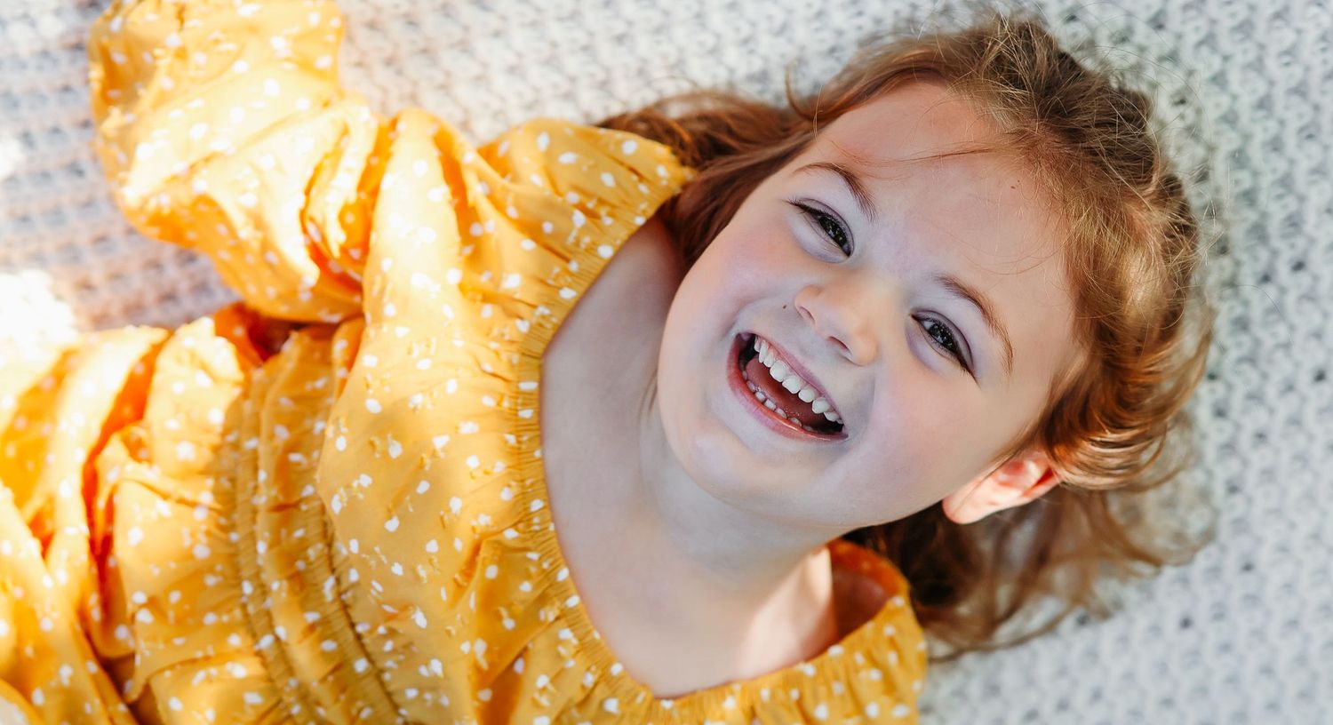 Joyful redhead girl in yellow polka dot dress laughing while lying on a white blanket.