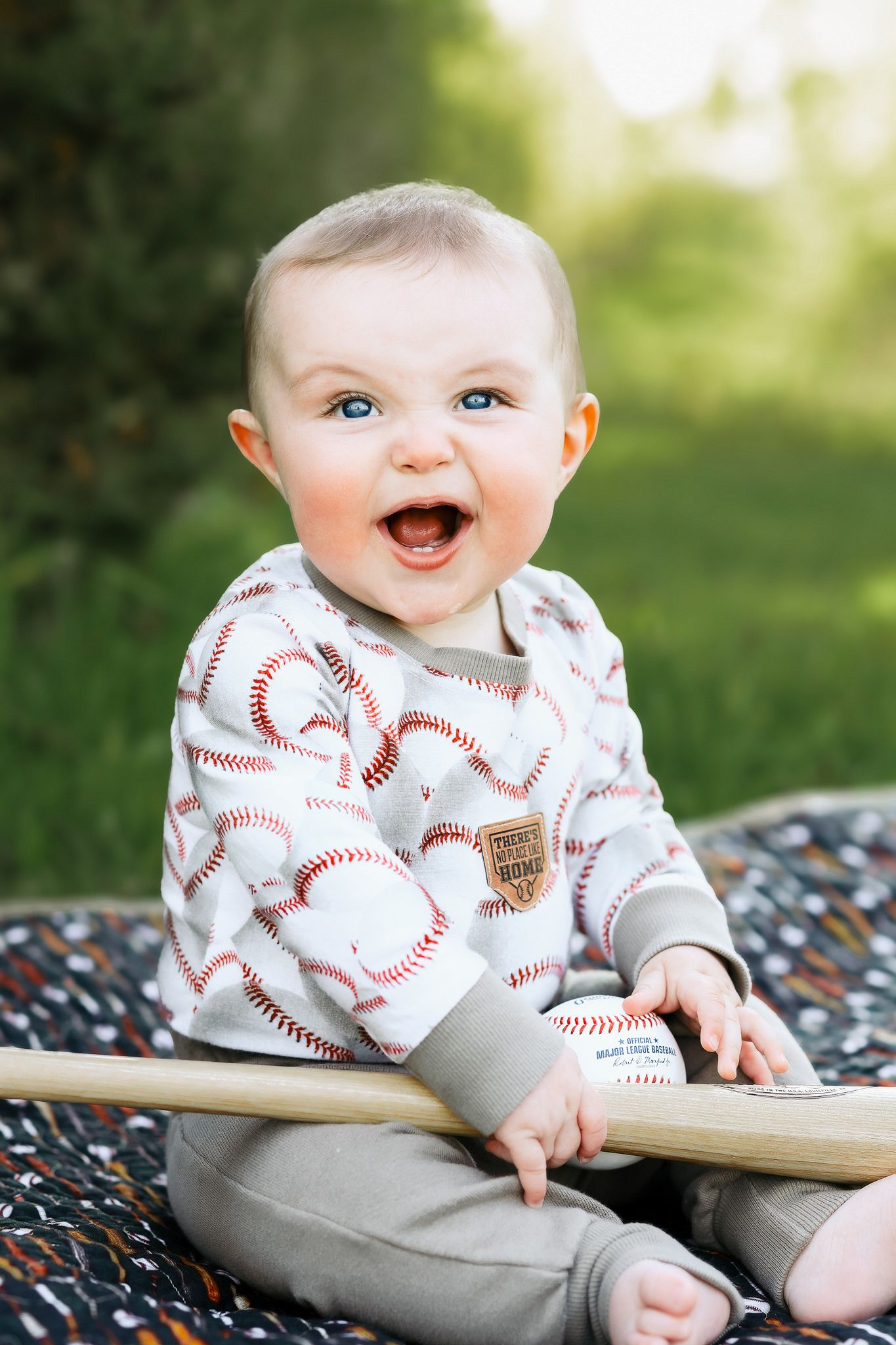 Happy baby boy in baseball-print hoodie laughing while holding a wooden bat outdoors.