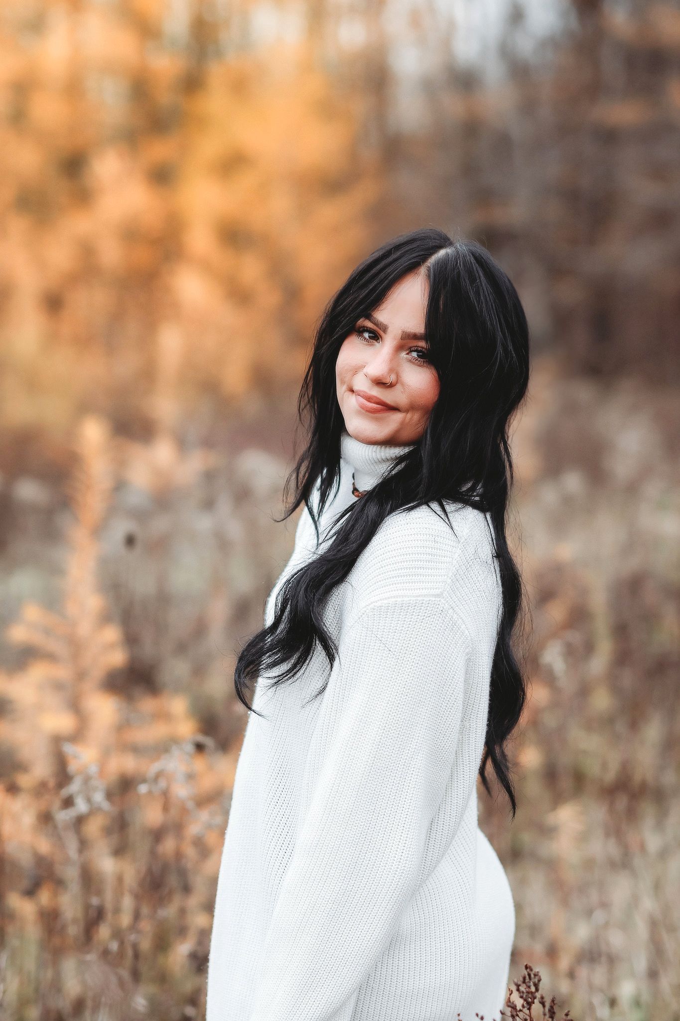 Beautiful dark-haired woman in white turtleneck sweater smiling over shoulder in fall outdoor setting.