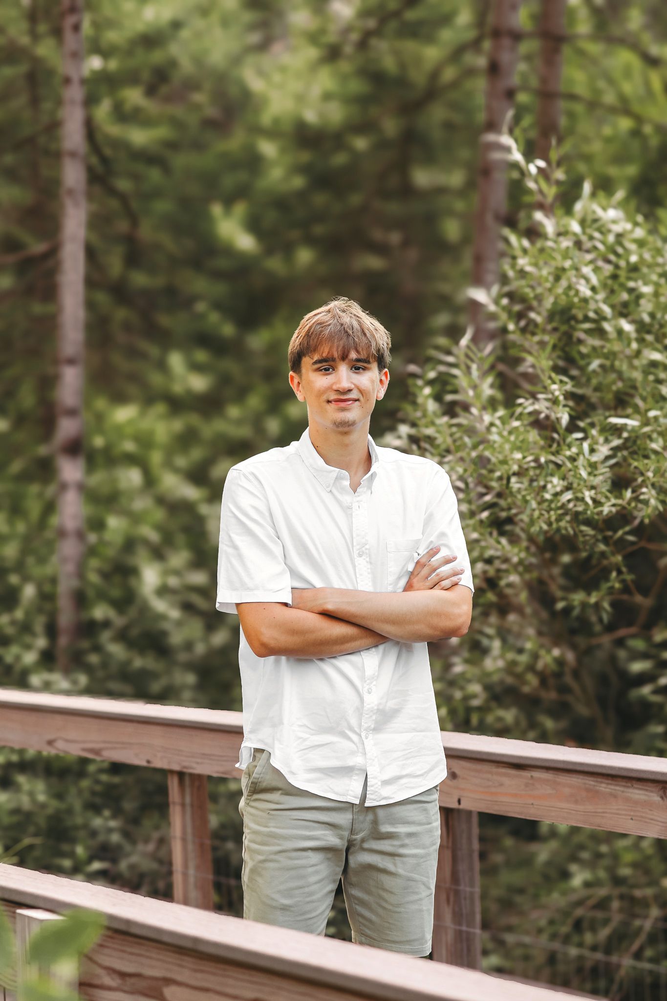 Smiling young man in white shirt with arms crossed posing on a wooden bridge in a forest.