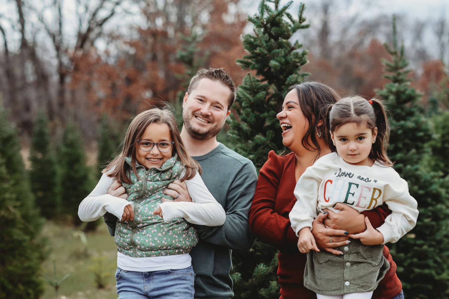 Joyful family of four poses and laughs together among Christmas trees outdoors.