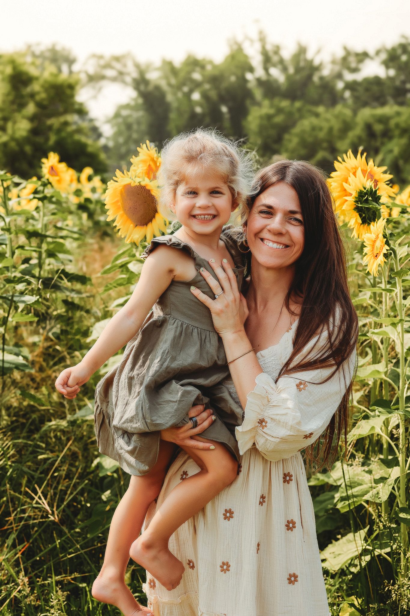 Smiling mother holding barefoot daughter with sunflowers in hair in a bright sunflower field.