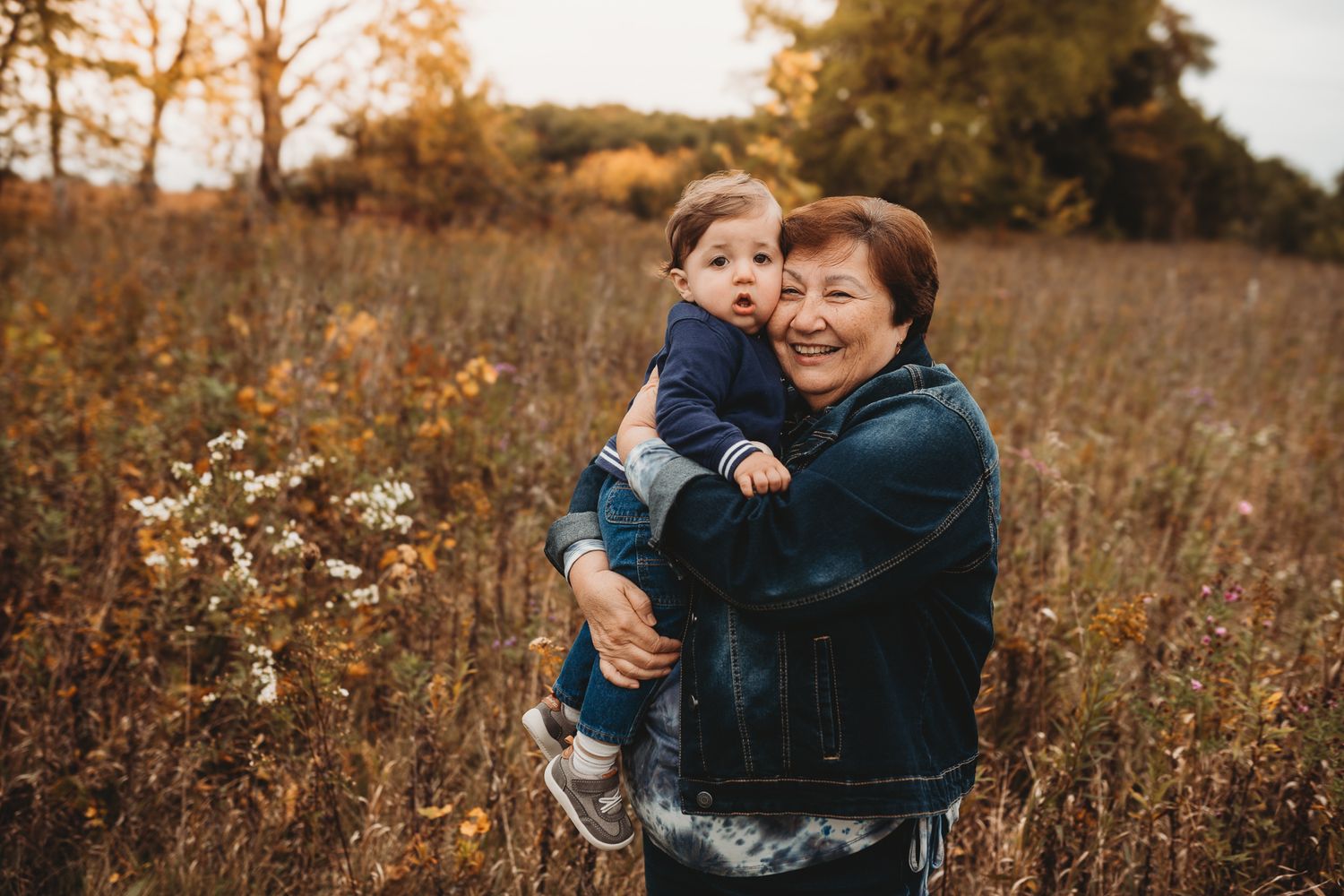 Grandmother smiles while holding a toddler in a golden autumn field.