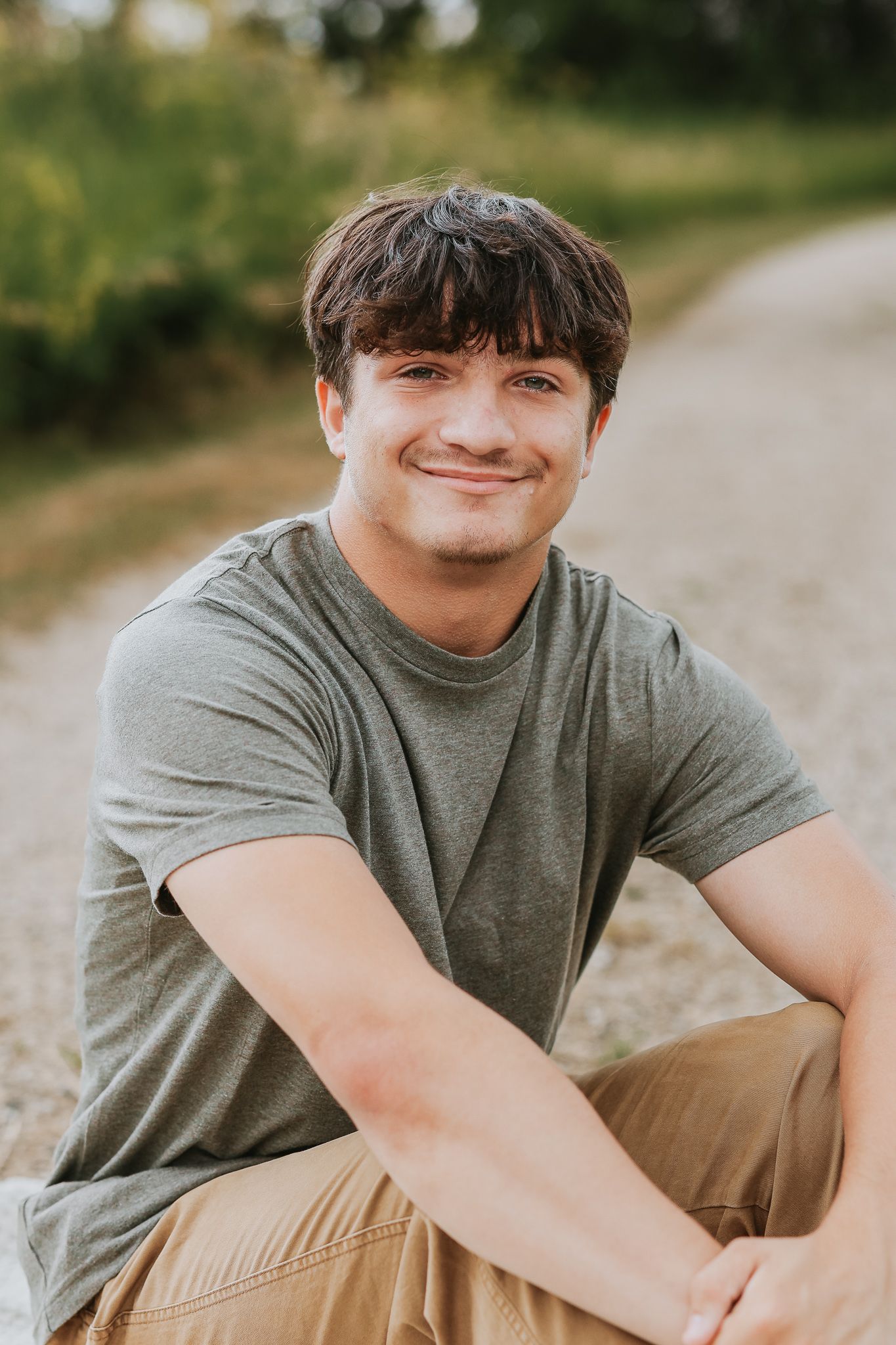 Young man in gray t-shirt sitting on gravel path smiling at camera outdoors.