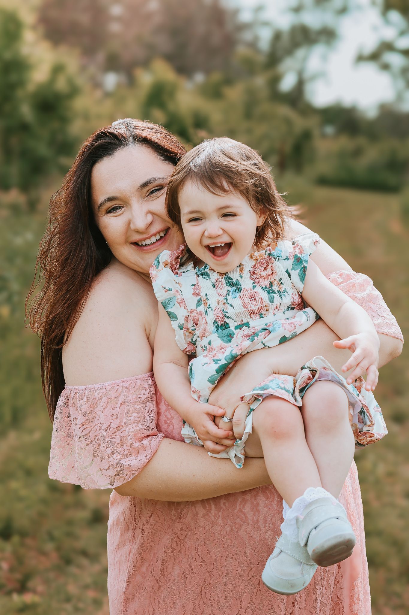 Smiling mother in pink off-shoulder dress holding laughing toddler girl in floral dress outdoors in nature.