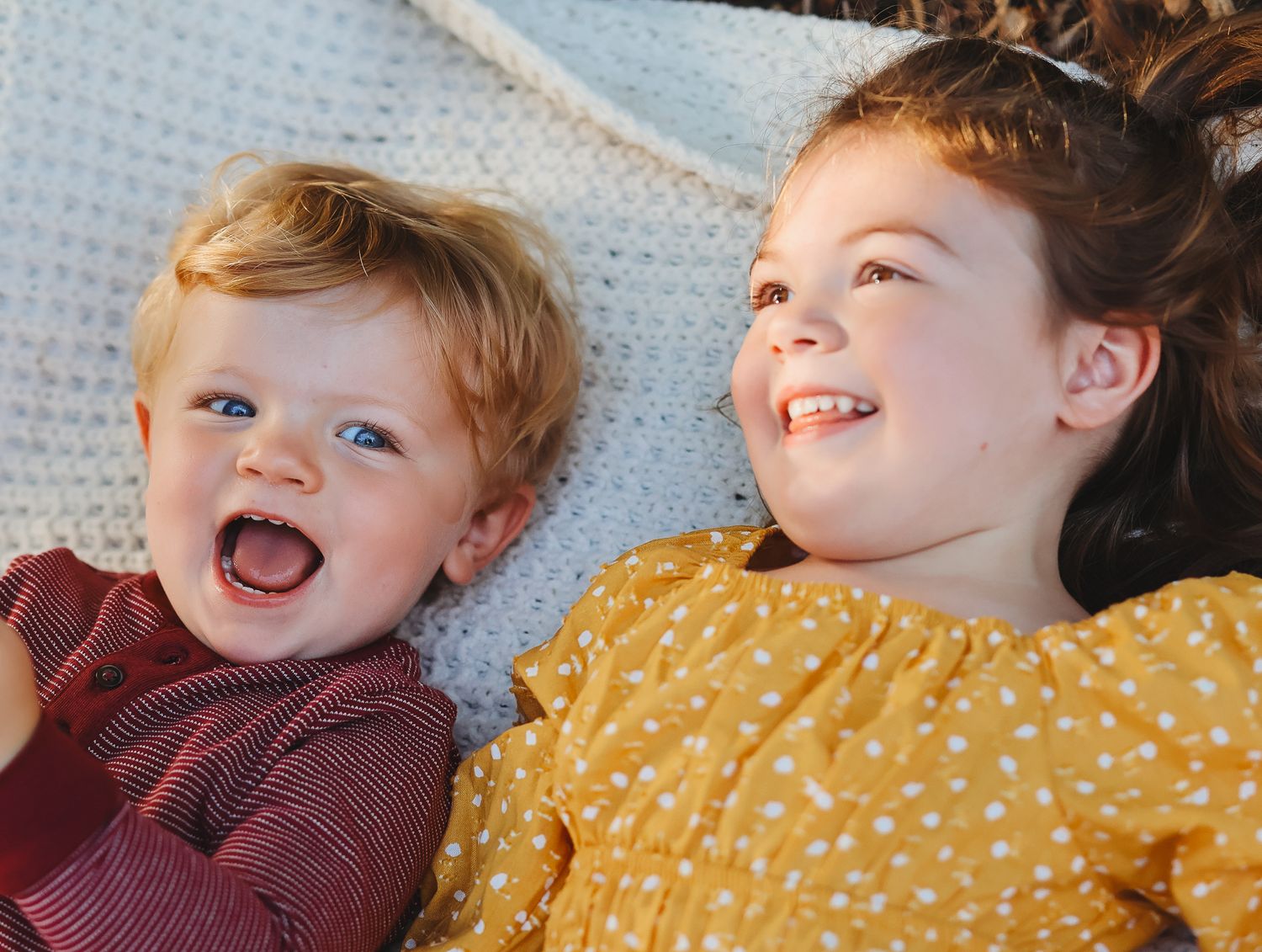 Two young siblings laughing and lying together on a white knit blanket outdoors.